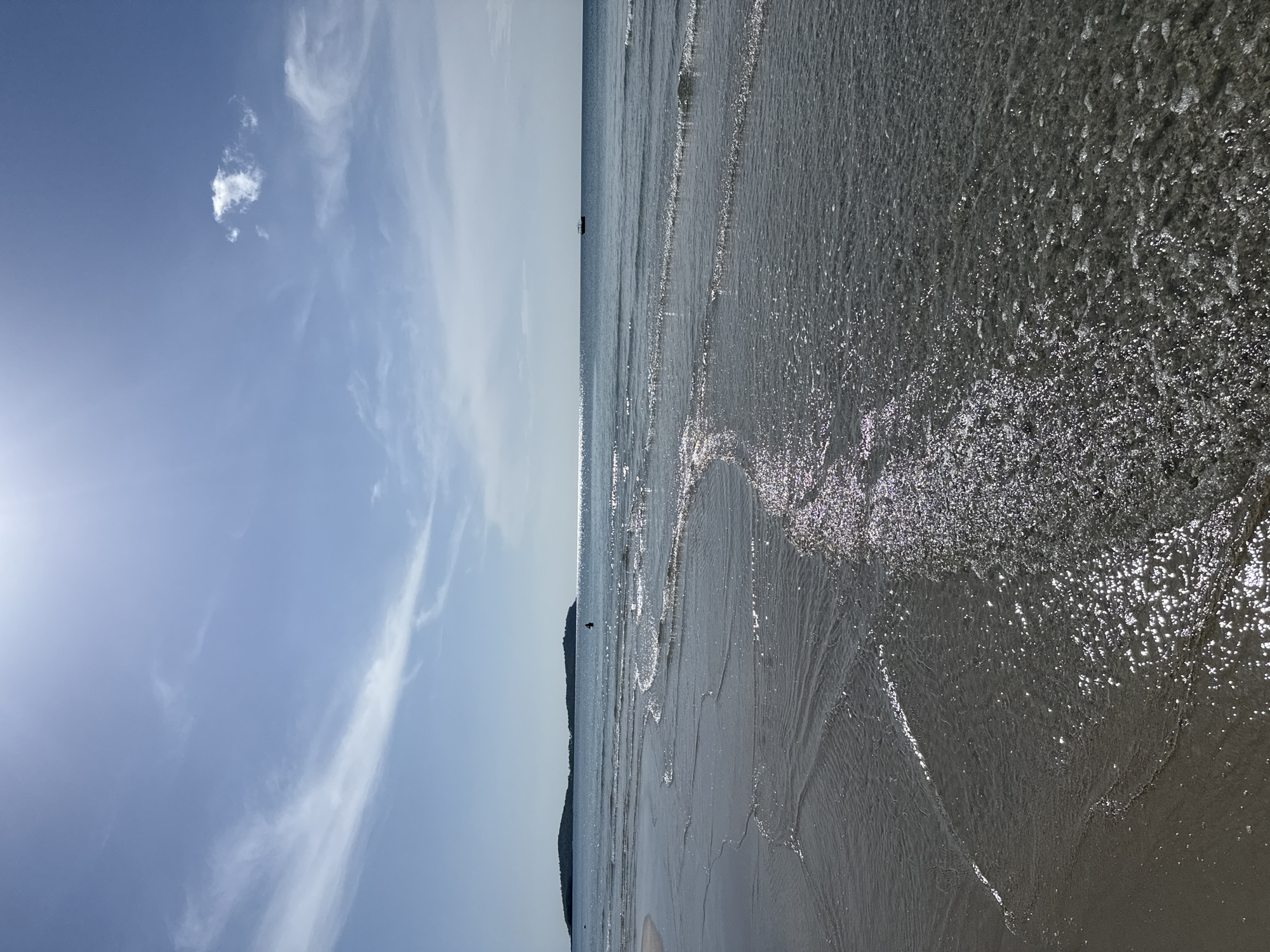 A vertical, eye-level shot captures a beach in the city of Ko Samui District, Thailand, under a bright, sunny sky. The foreground is dominated by shallow, receding waves on a sandy beach. The water, clear and gently rippling, reflects the sunlight, creating a shimmering effect. In the mid-ground, a person is visible standing in the water, appearing as a dark silhouette against the bright sea. Further out, a small boat floats on the horizon. Two small islands, covered in dense foliage, are visible in the distance. The sky is a clear blue, with wispy white clouds and the sun creating a lens flare in the upper portion of the image. The overall scene depicts a tranquil beach environment on a sunny day.