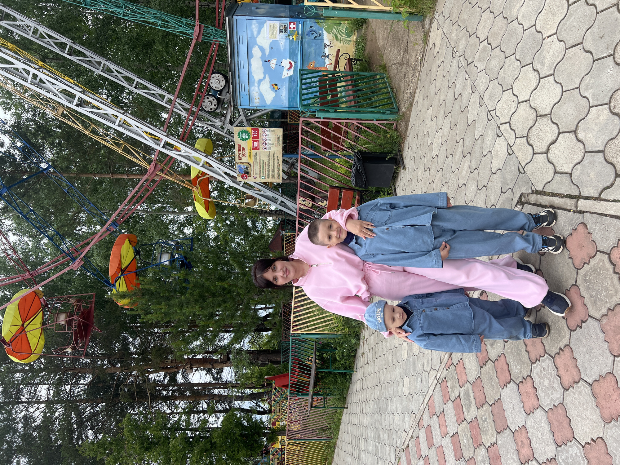 This image captures a family group in an amusement park located in Chaikovsky, Russia. In the foreground, an adult female stands between two young boys on a paved walkway featuring interlocking grey and reddish-brown pavers. The female, with dark hair, wears a light pink tracksuit with a zipper at the collar and dark sneakers with white soles. Her right arm is around the older boy's shoulder, and her left hand rests on the younger boy's left shoulder. The older boy, positioned to her right, wears a light blue denim jacket and matching pants, with a dark shirt underneath and dark sneakers with white soles. He has short hair and a slight smile. The younger boy, to her left, wears a similar light blue denim jacket and pants, a dark undershirt, and a blue cap with "BOSTON" embroidered on it, along with dark shoes. He is looking downwards.
Behind the group, a vibrant amusement park environment is visible. A large, multi-colored ferris wheel dominates the background, with structural elements in red, yellow, blue, white, and green. Several gondolas with yellow and orange parasol-like covers are attached to the wheel. At the base of the ferris wheel, a control booth or structure is visible, featuring a large white sign with Cyrillic text indicating "Panoramic View" (КРУГОВОЙ ОБЗОР), "One Circle" (ОДИН КРУГ), and prices "180" and "130" (likely rubles). Adjacent to this, a blue rectangular structure is painted with a seaside scene featuring a sailboat, fish, and seabirds, and includes a small white sign with "59" and a red cross symbol.
Various colorful fences, primarily pink, yellow, and green, delineate different areas of the park, with red and green benches visible behind them. A black trash receptacle is located near a red bench. The background is filled with numerous tall pine and deciduous trees under an overcast sky, suggesting a park setting. The group is stationary, posing for a photograph.