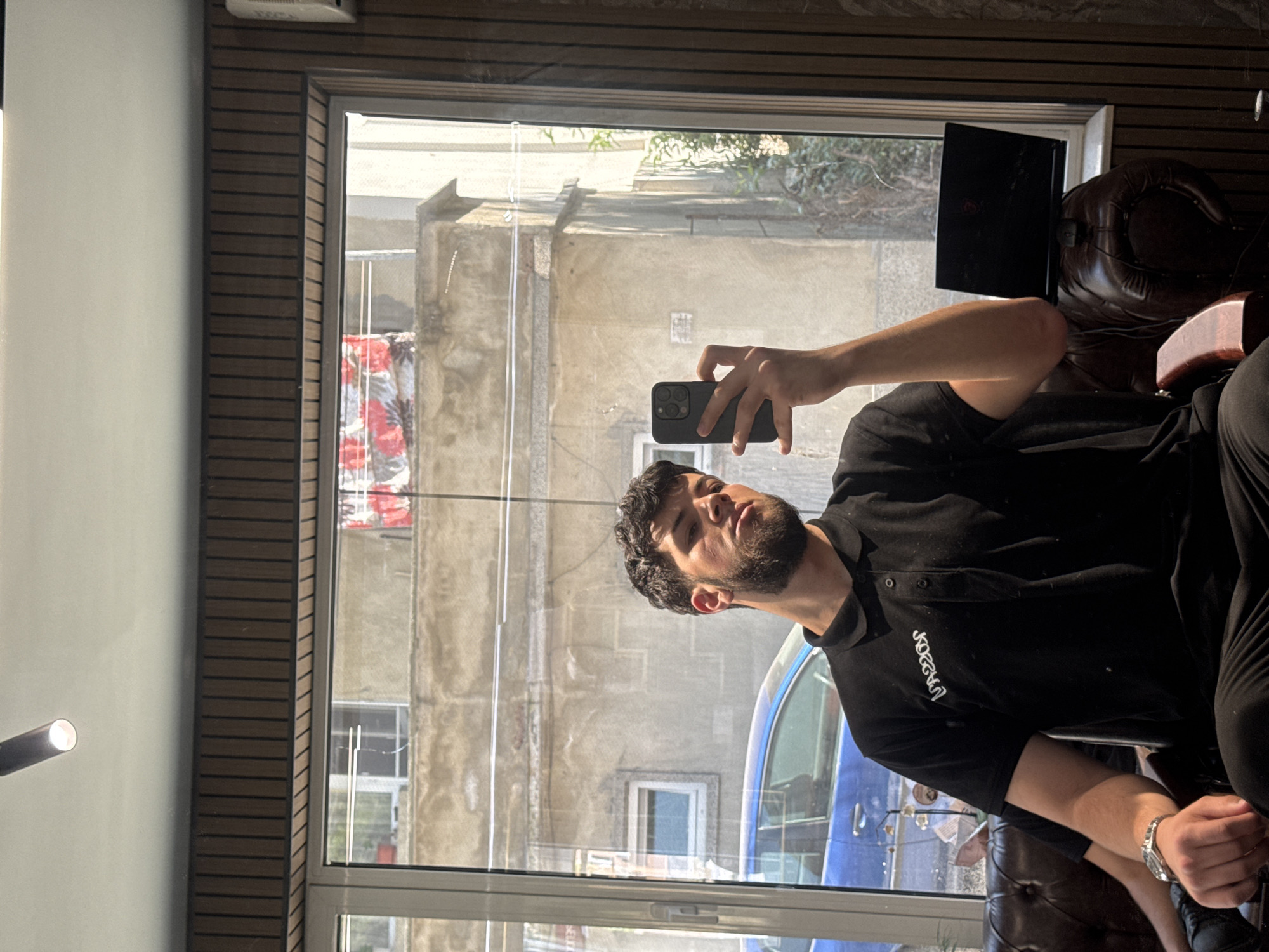 An image, originally rotated 90 degrees clockwise, depicts a young man sitting indoors next to a large window, appearing to take a selfie or video with his smartphone. The man has dark, curly hair and a beard, and is wearing a black polo shirt with "JOKERS" embroidered in white on the left chest, and dark pants. A silver watch is visible on his left wrist. He holds a black smartphone with a triple-lens camera in his right hand, angled towards himself.
The indoor setting features a white wall on the left, contrasting with a section of dark brown wooden slat paneling to the right, framing the window. A modern tubular light fixture is partially visible on the far left. Part of a brown leather armchair is seen to the man's right, and a black laptop rests on what appears to be another chair or surface behind him, near the window.
Through the window, an urban street scene is visible. The view shows weathered concrete or stucco buildings with visible windows, suggesting an older architectural style. A blue car is partially reflected or parked outside, catching the sunlight. Some sparse greenery is also visible, along with a colorful reflection or banner of red and white patterns across the street. The bright natural light streaming through the window indicates it is daytime, casting strong illumination into the room. Given the context of Varna, Bulgaria, the scene captures a typical moment in an urban environment.