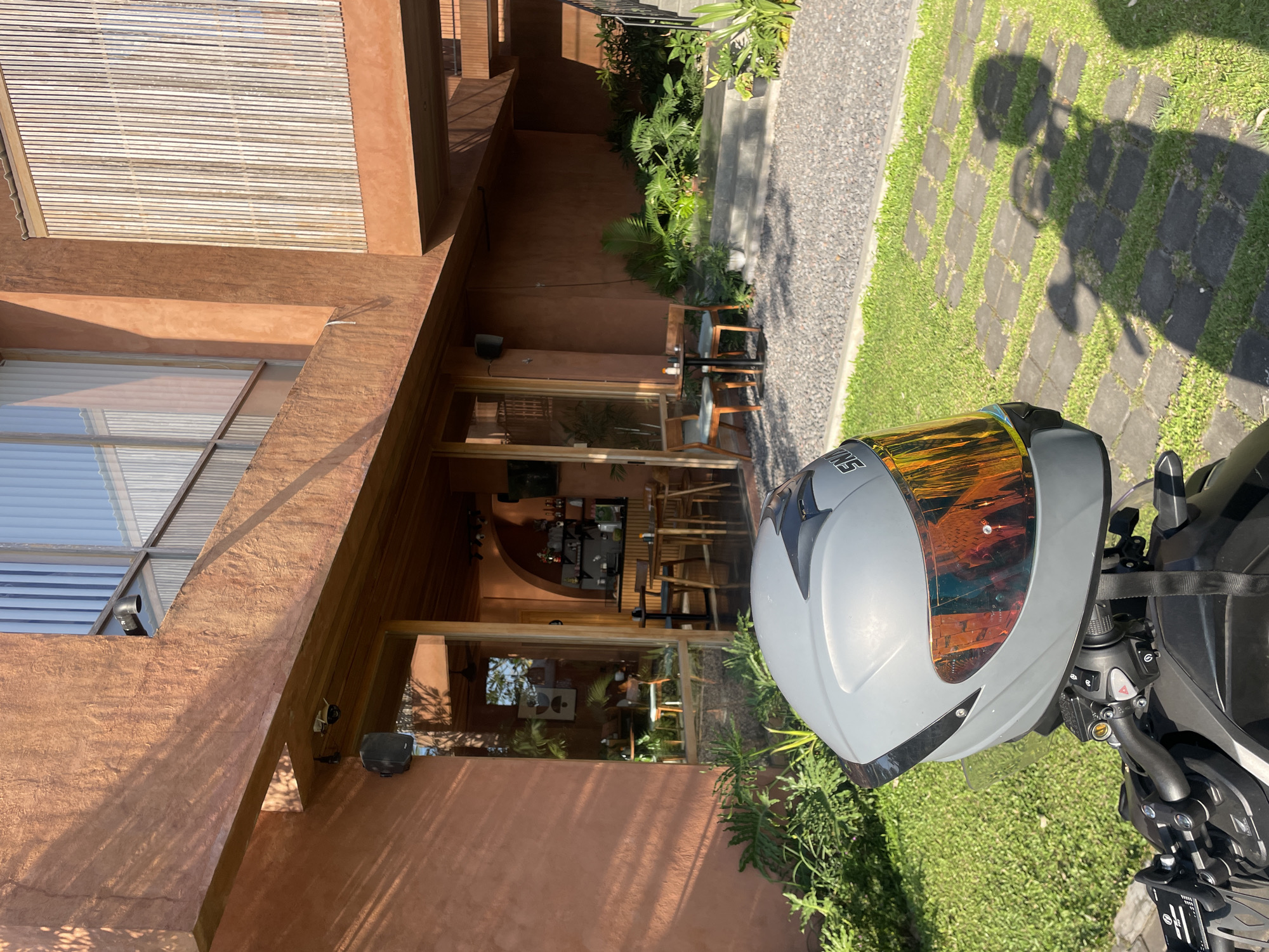 A motorcycle helmet with a reflective visor rests on the handlebars of a motorcycle parked on a stone path in front of a modern building with terracotta-colored walls. The building has large windows, some with vertical blinds, and wooden accents. Green grass and lush plants surround the area. The scene appears to be outdoors, possibly a cafe or restaurant in Kediri, Indonesia, given the warm lighting and tropical vegetation. The sun casts shadows on the ground, suggesting it is daytime.