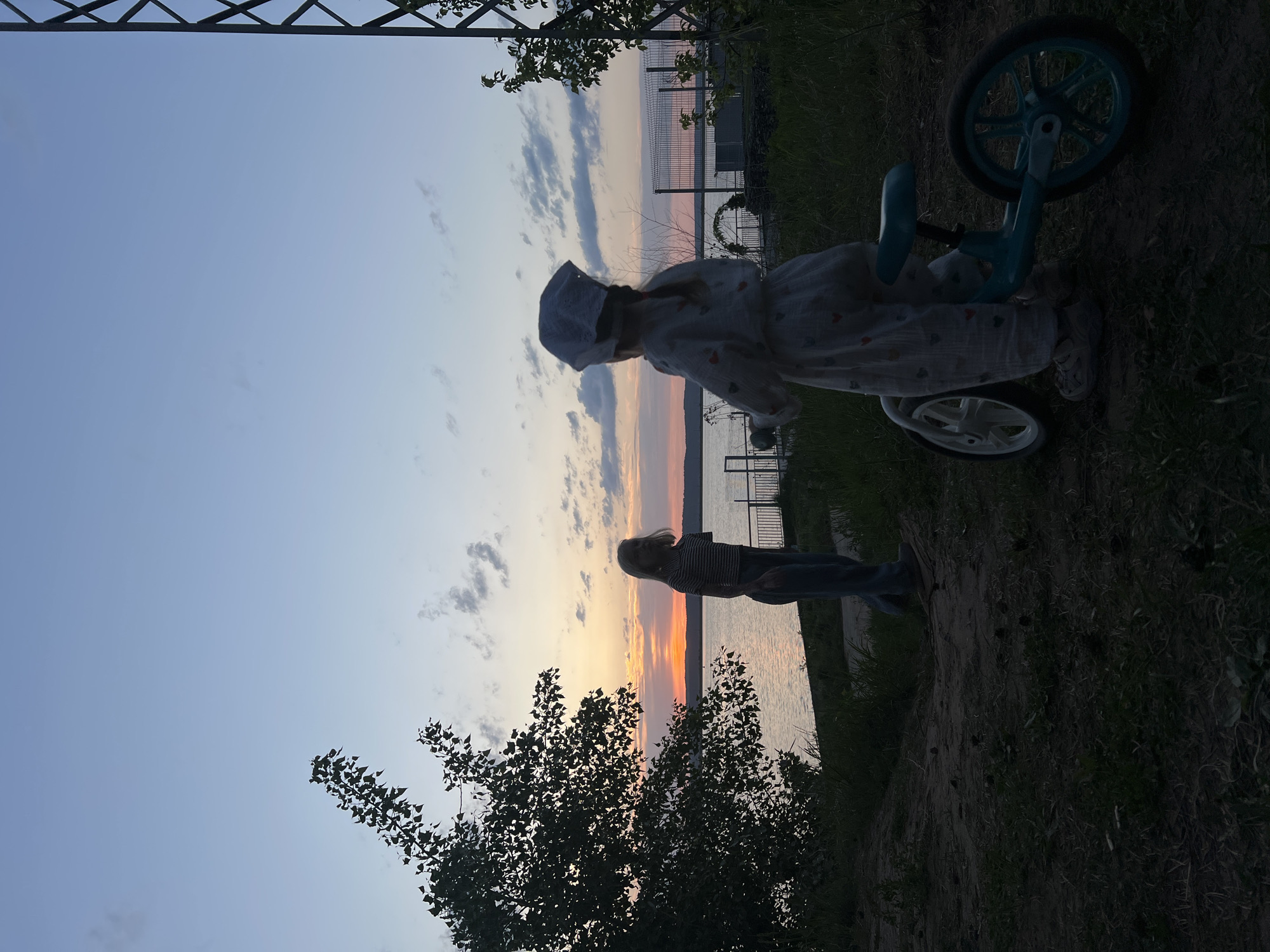 The image depicts a lakeside scene at dusk in Chaikovsky, Russia. The sky exhibits a gradient of colors, transitioning from blue to orange and pink near the horizon. A body of water reflects the sky's colors. Two figures are present: one is a woman, silhouetted against the sunset, the other is a child near a tricycle. Vegetation is visible, including trees and grass. A fence and a structure can be seen in the distance, suggesting an urban area.