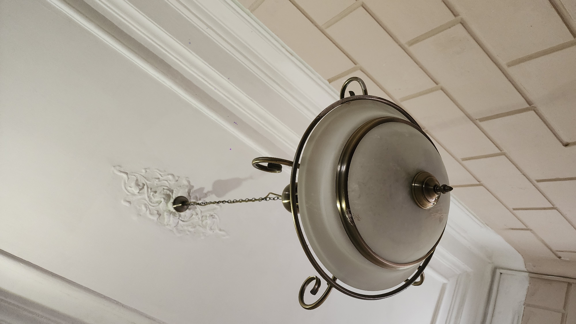 The image shows a low-angle view of a ceiling lamp in what appears to be an interior space. The lamp is round, with a frosted white glass shade encased in a dark metal frame featuring decorative curves. It hangs from the ceiling via a chain attached to a decorative medallion. The ceiling itself is white with ornate molding. In the upper right corner, a section of tiled wall is visible, indicating the proximity of a wall or potentially a backsplash if this is a kitchen or bathroom. Given the architectural details, it may be possible that the scene is a building in Moscow, Russia, as Moscow has a large number of buildings with ornamented stucco or plaster ceilings, and pendant lights with frosted glass shades in bronze or antique brass fittings. Without further information, it's impossible to confirm the exact location.