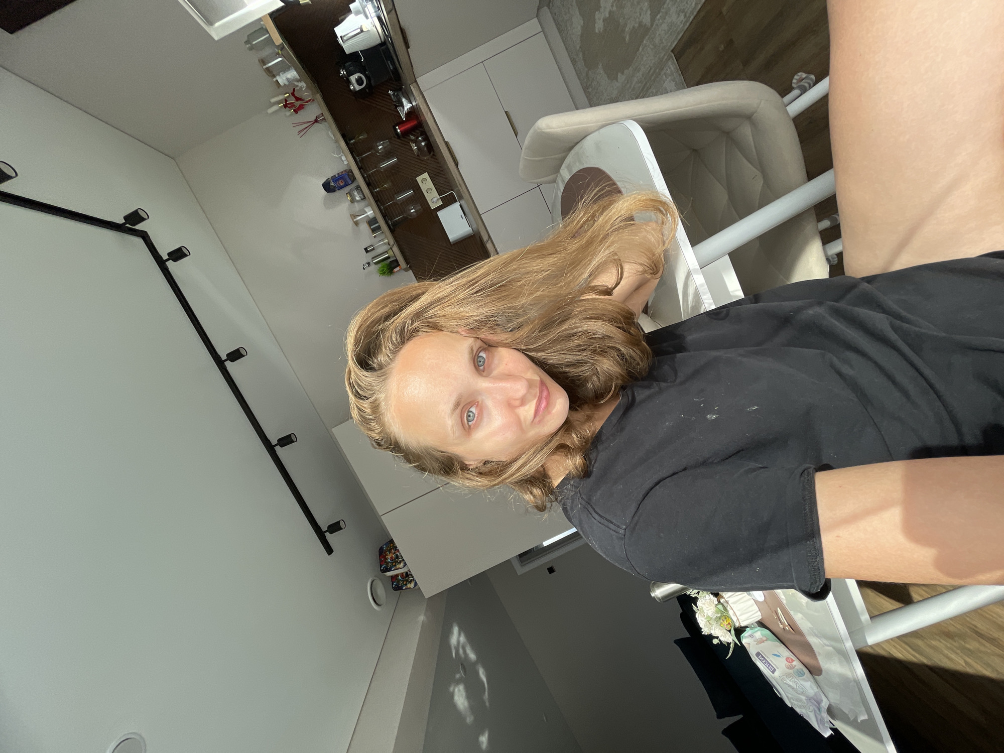 A medium shot captures a woman from the chest up, seated at a white table with light wooden legs and a cream-colored chair. She has long, wavy blonde hair that falls over her right shoulder and is resting her head on her hand. Her eyes are blue and she is looking directly at the camera. She is wearing a plain black t-shirt. To her left, on the table, is a small bouquet of white flowers in a vase and a package of wet wipes. The background reveals a modern kitchen with white cabinets, a wooden backsplash, and a shelf stocked with jars and bottles. Above, black track lighting illuminates the space. Sunlight streams in from the left, casting shadows on the wall and ceiling, suggesting a residential interior, likely a dining area or kitchen in a home, potentially in Podshivalovo, Russia.