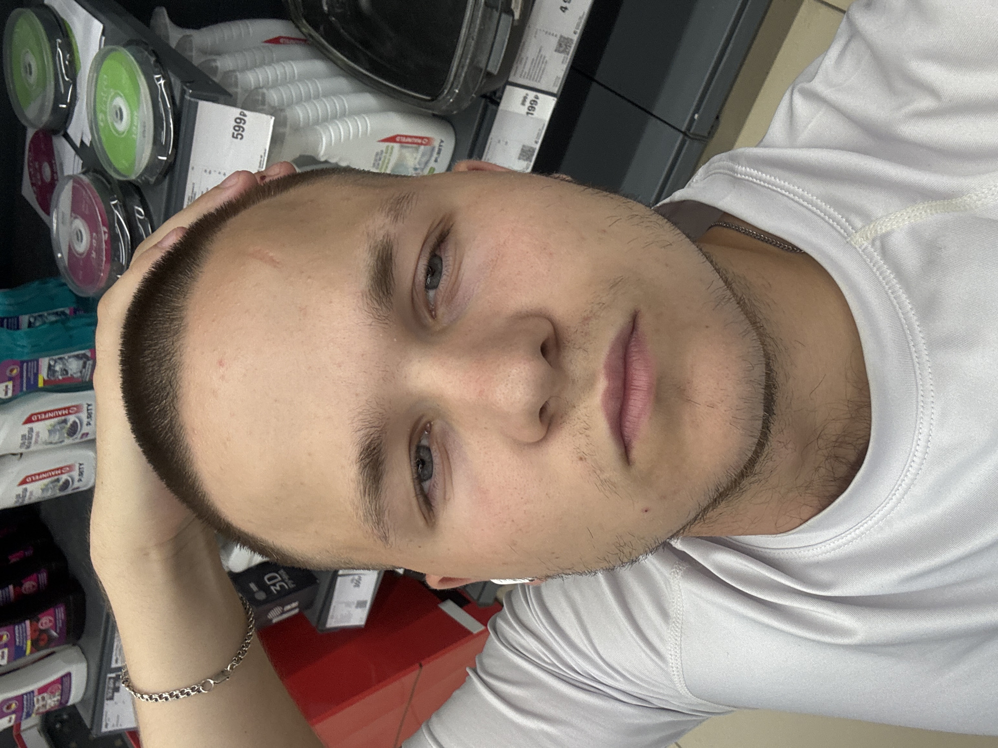 A young man with a buzz cut and light stubble is holding his head with his left arm. He is wearing a light grey long-sleeved t-shirt. In the background, there are shelves with various products, including what appear to be cleaning supplies and compact discs. Price tags are visible on the shelves, with one reading "599p". The overall setting suggests a retail environment, potentially a supermarket or discount store. There is no visual information that definitively places the image in Ishim, Russia.