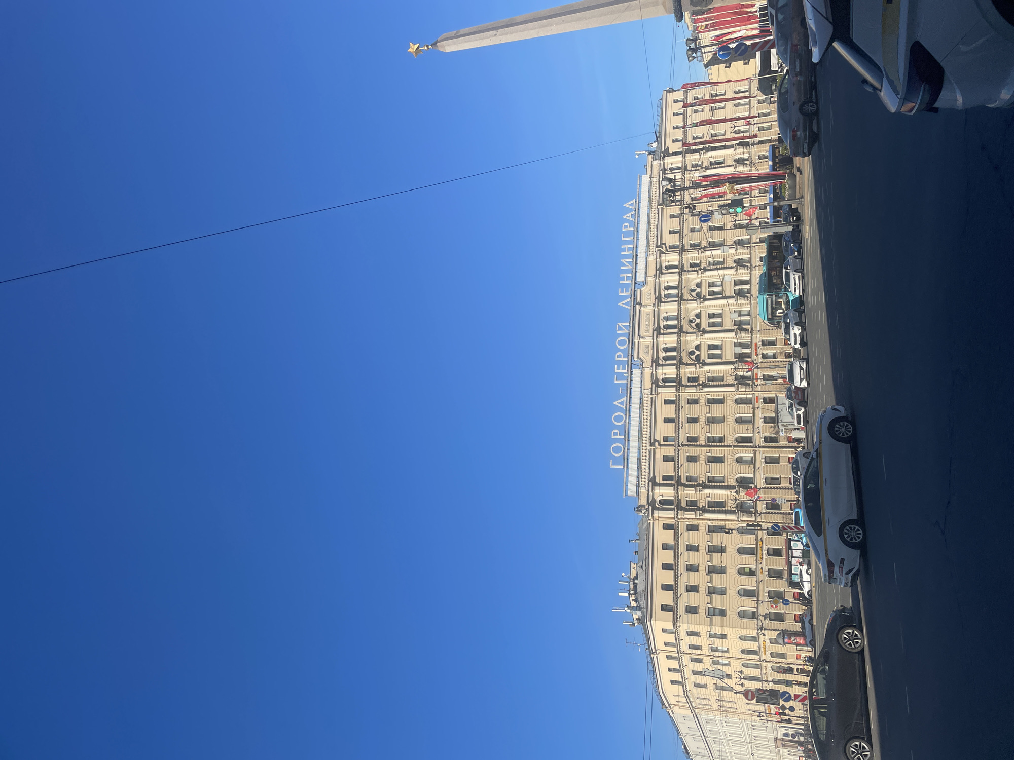This media file captures a vibrant urban street scene in Saint Petersburg, Russia, under a clear, bright blue sky. The photo is taken during the daytime, with ample sunlight illuminating the buildings and street.
On the right side of the frame, a large, multi-story building with ornate historical architecture dominates, characterized by numerous arched windows and a light beige facade. White letters spell out "ГОРОД-ГЕРОЙ ЛЕНИНГРАД" (HERO CITY LENINGRAD) along its upper part, serving as a significant historical and geographical marker. Several red banners or flags hang vertically from the building, adding a splash of color.
To the left, a tall, light-colored obelisk or monument rises prominently into the sky, topped with a distinctive golden star. A thin black overhead wire stretches across the blue sky, adding a subtle linear element to the composition.
The street below the building is busy with vehicles. Several cars are visible, including a white car in the foreground on the right and other parked cars along the curb. Traffic signs are also visible on the street. The overall scene depicts a bustling city environment on a sunny day, with the historical text on the building firmly placing the location in Saint Petersburg, formerly known as Leningrad. No people are distinctly visible.