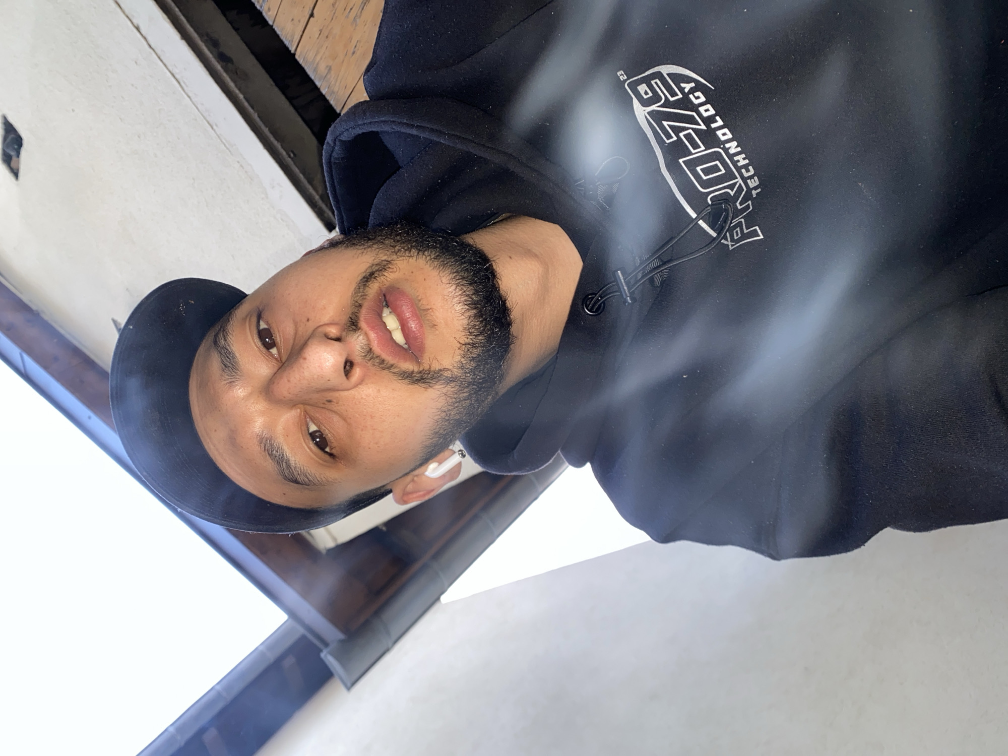 A man wearing a black hoodie with the "PRO-ZE TECHNOLOGY" logo, a black cap, and an earpiece is posing for a selfie. The man has a short beard and light skin. He is looking down at the camera. A glimpse of the building exterior shows a wooden board at the top and white walls and a dark roof on the left. The image was likely taken in Schiltigheim, France.