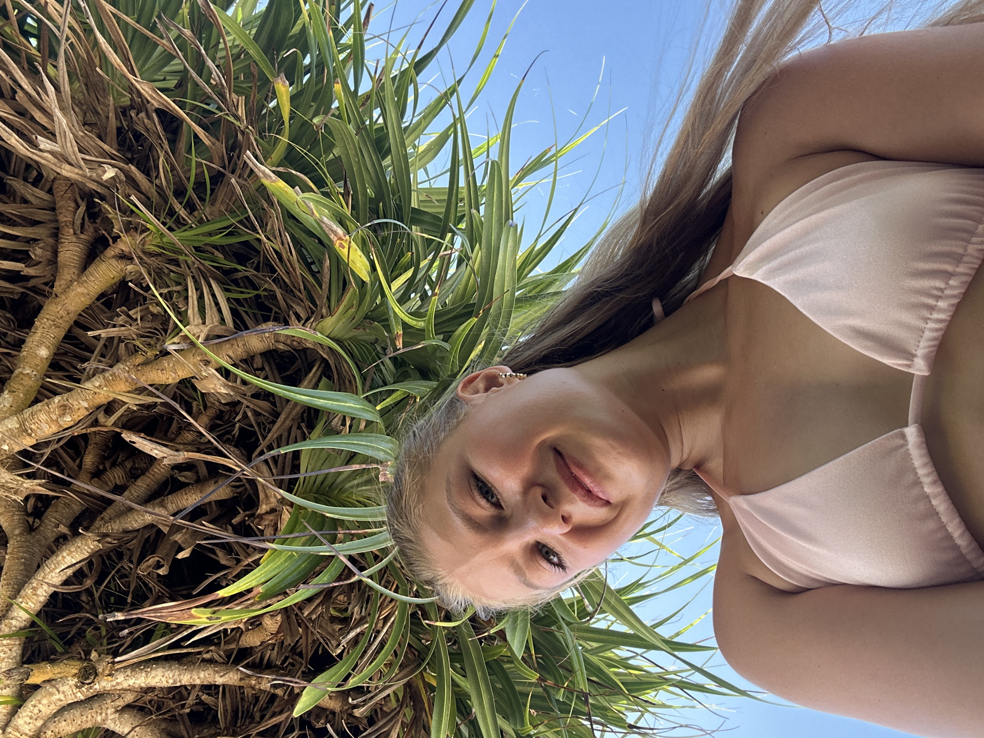 A woman in a pink bikini is standing near a pandanus tree in the Kuta Selatan region of Indonesia. She is positioned beneath the tree, looking down at the camera with a slight smile. The sky is visible in the upper right corner of the frame. The tree's exposed roots and green spiky leaves are visible.