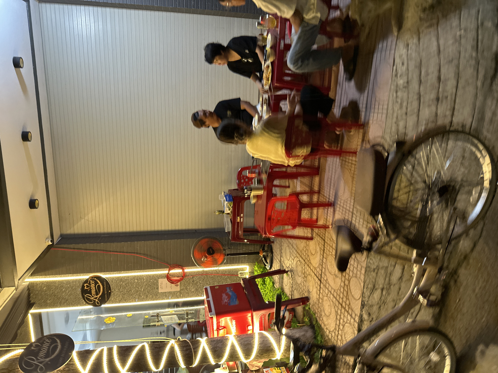 The image shows an outdoor dining area in Bac Nha Trang Ward, Vietnam. Patrons are seated at red plastic tables and chairs. A bicycle is parked to the right. A storefront with the sign "Romance" illuminated by string lights is visible. A red cooler and an orange fan are positioned near the storefront. The scene is lit by streetlights and restaurant lighting.