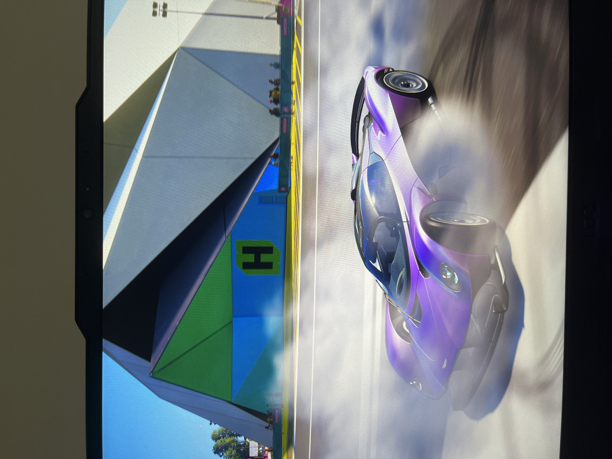 The image shows a video game scene with a purple car drifting on a track next to a building with a green and blue "H" on it. The building is white with geometric shapes. There are people standing near the building. The environment appears to be sunny. The location is not provided in the image, but a city named "Unknown, Unknown" is mentioned.
