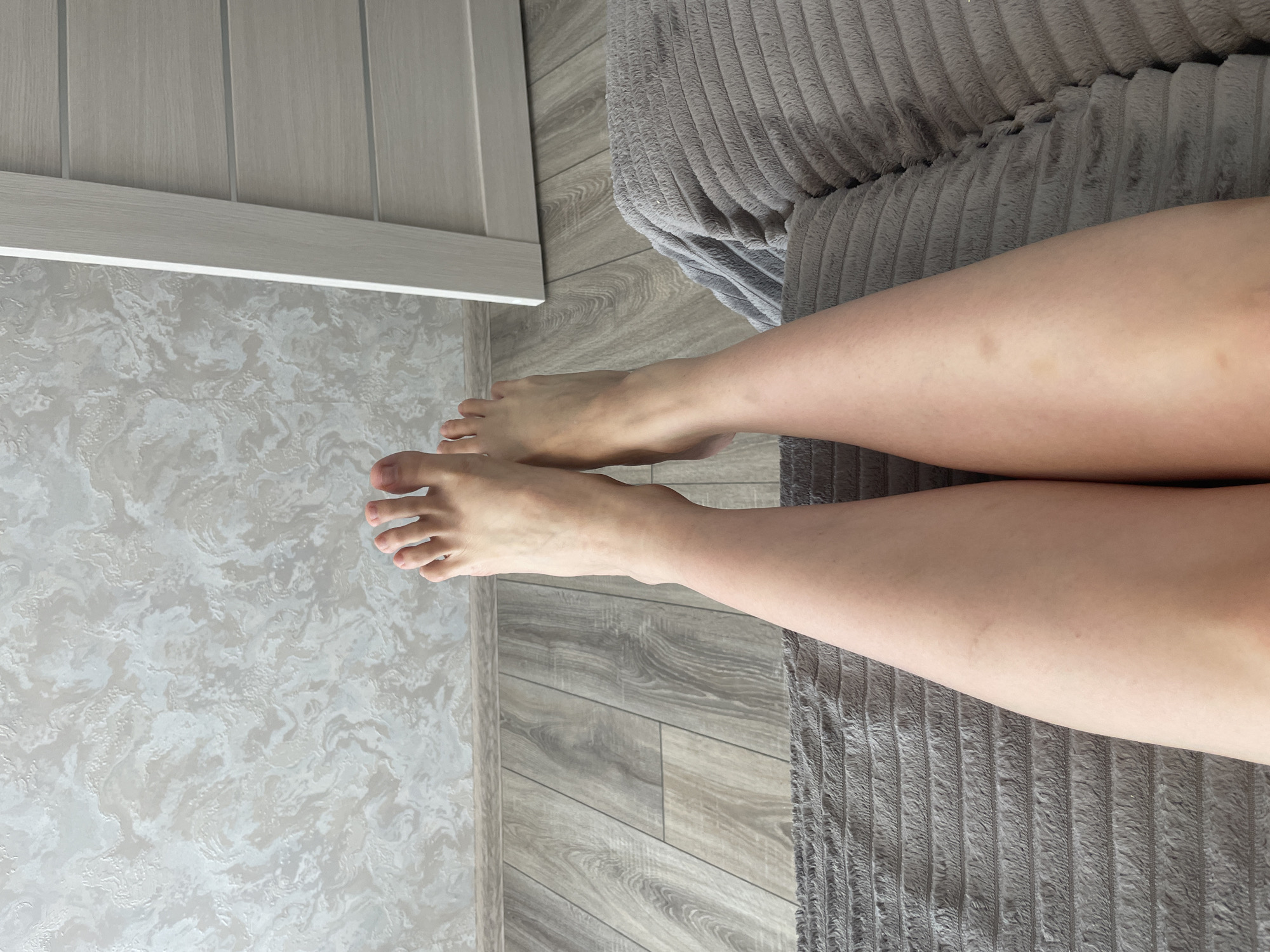 The image displays a close-up, low-angle perspective of a person's legs and feet, resting on a textured gray blanket or cushion. The legs are bare, and the feet are visible with toes spread. The background consists of a wall with textured wallpaper and a wooden floor. To the right, a portion of a light-colored door is visible. The scene appears to be indoors, likely in a residential setting. There is no indication of the location being Stavropol, Russia.
