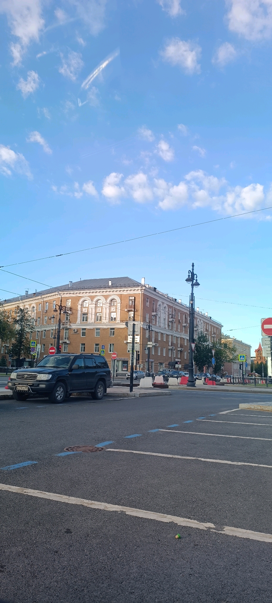 The image shows a wide street in front of a large, multi-story apartment building with a red-brown facade and arched windows. The building is characteristic of Stalinist architecture. A dark-colored SUV is parked on the side of the road. There are traffic lights, street lamps, and traffic signs visible. The sky is blue with some white clouds. The street appears to be an intersection, with road markings and a manhole cover visible on the asphalt. A small green and white object, possibly a shuttlecock, is lying on the road in the foreground. The presence of Cyrillic text on a sign on the building suggests the location is in Russia. While the exact location within Perm is not specified by any visible signage, the architectural style is consistent with buildings found in cities across Russia, including Perm.