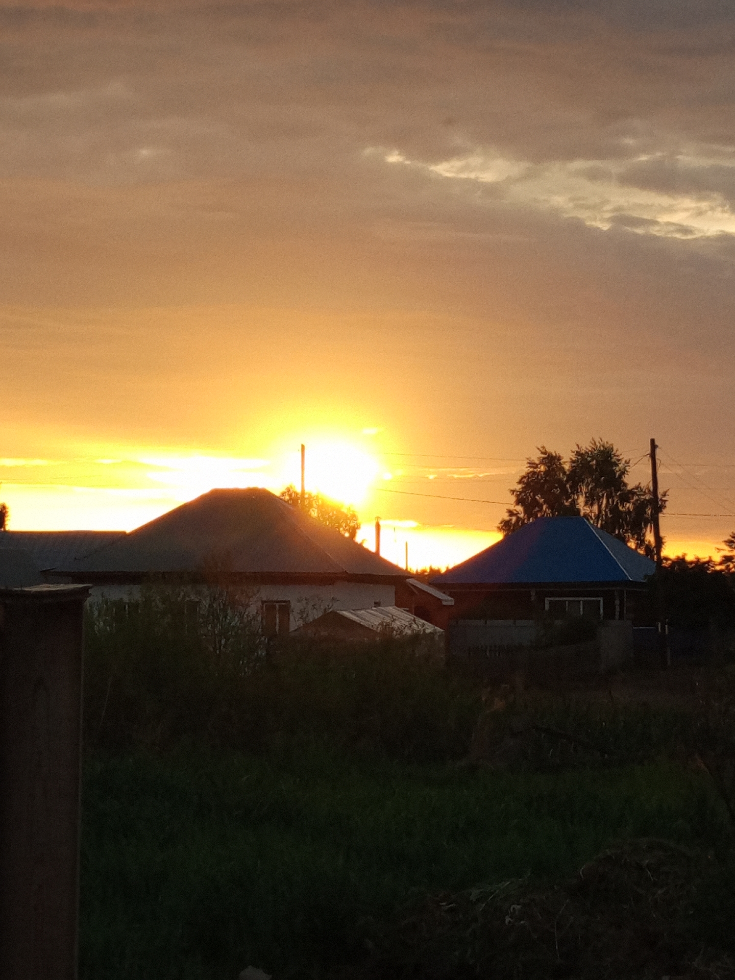 The image depicts a rural sunset scene in Pankrushikha, Russia. The sun, a bright orb of light, is setting near the horizon, casting a warm, orange glow across the sky and illuminating the clouds. In the foreground, silhouetted against the vibrant sky, are several residential structures. One prominent building has a dark, peaked roof, while another nearby structure features a blue roof. Trees and vegetation are visible in the mid-ground and foreground, rendered in dark tones due to the low light conditions. A wooden pole, possibly for utility lines, stands to the right of the frame. The overall impression is one of tranquility and the end of a day in a rural setting.