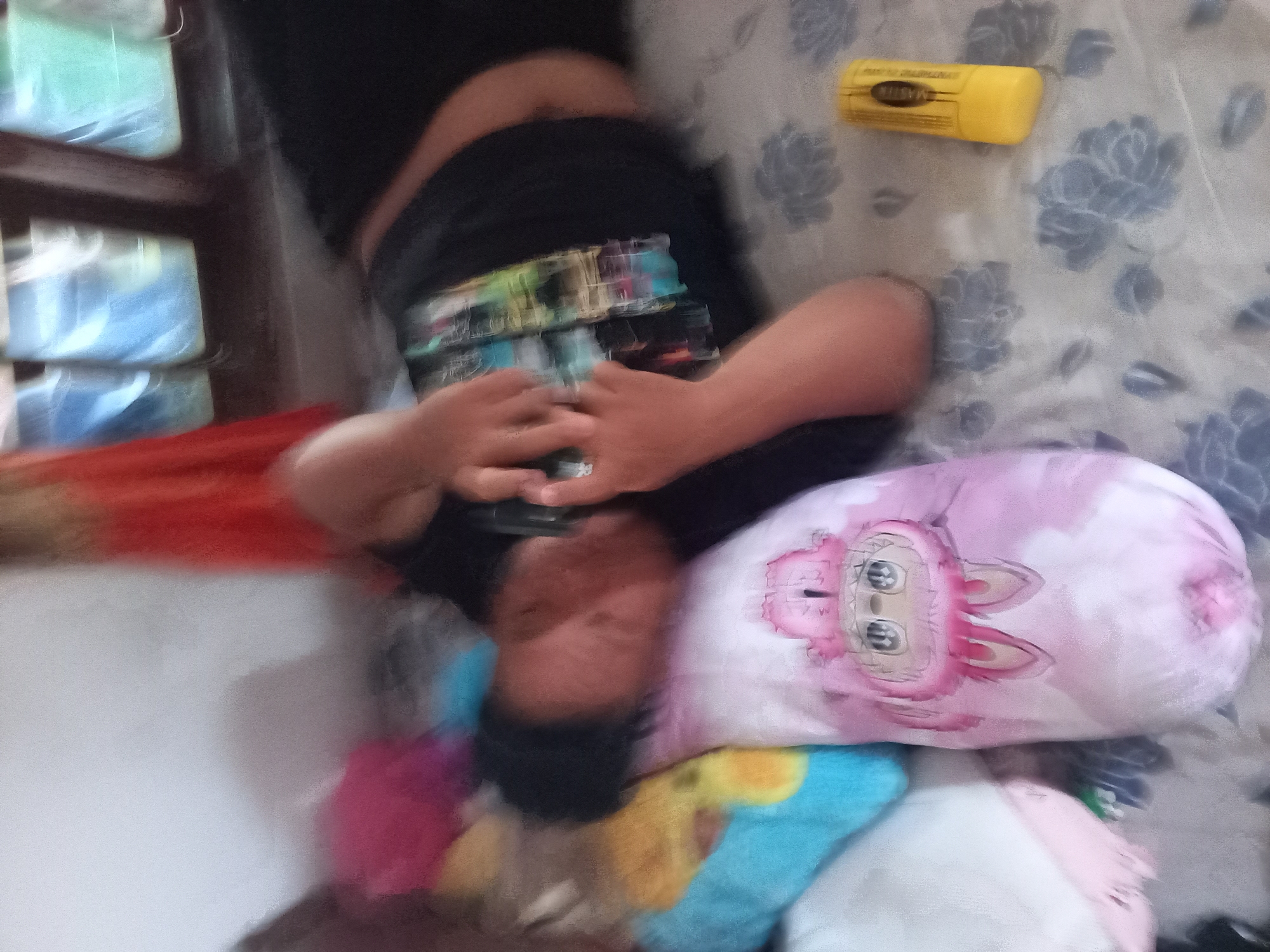 A person is lying on their side on a patterned bed, holding a phone to their ear. The person is wearing a black shirt with a colorful pattern on it. The bed is covered with a light-colored sheet with a floral pattern. There are several pillows on the bed, including a pink pillow with a cartoon character on it. To the right of the person, there is a yellow object, possibly a tube of sunscreen or lip balm. In the background, there is a window with blinds, and a red curtain is partially visible. The overall lighting suggests that the image was taken indoors during the daytime. The location is described as Kebon Agung, Indonesia.