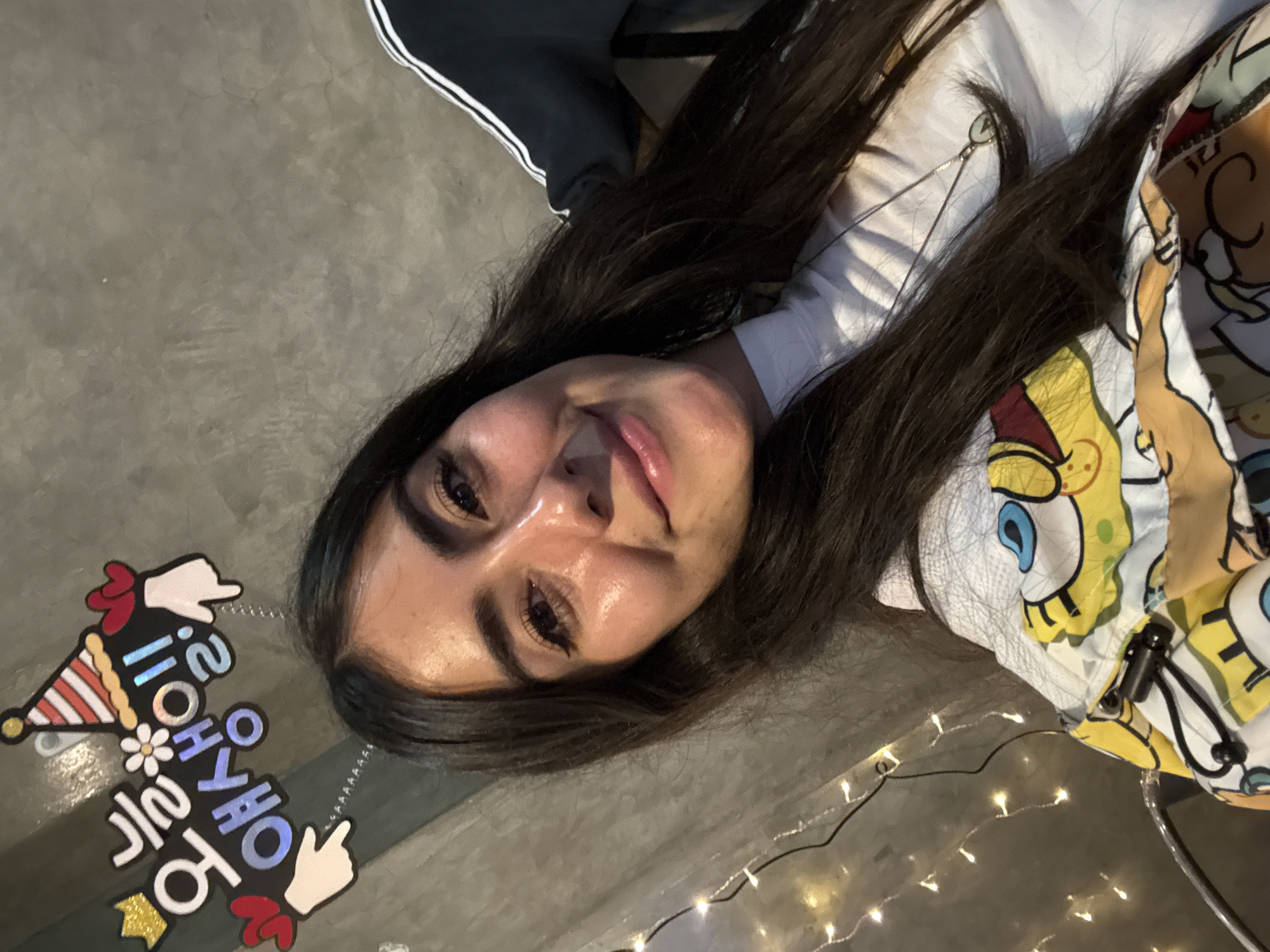 A person with dark hair lies on a gray surface, wearing a white shirt under a patterned jacket featuring SpongeBob SquarePants. Part of black pants with a white stripe is visible. A headband with Korean writing and festive decorations rests beside the person. String lights illuminate the area. The location is the city of Puebla, Mexico.
