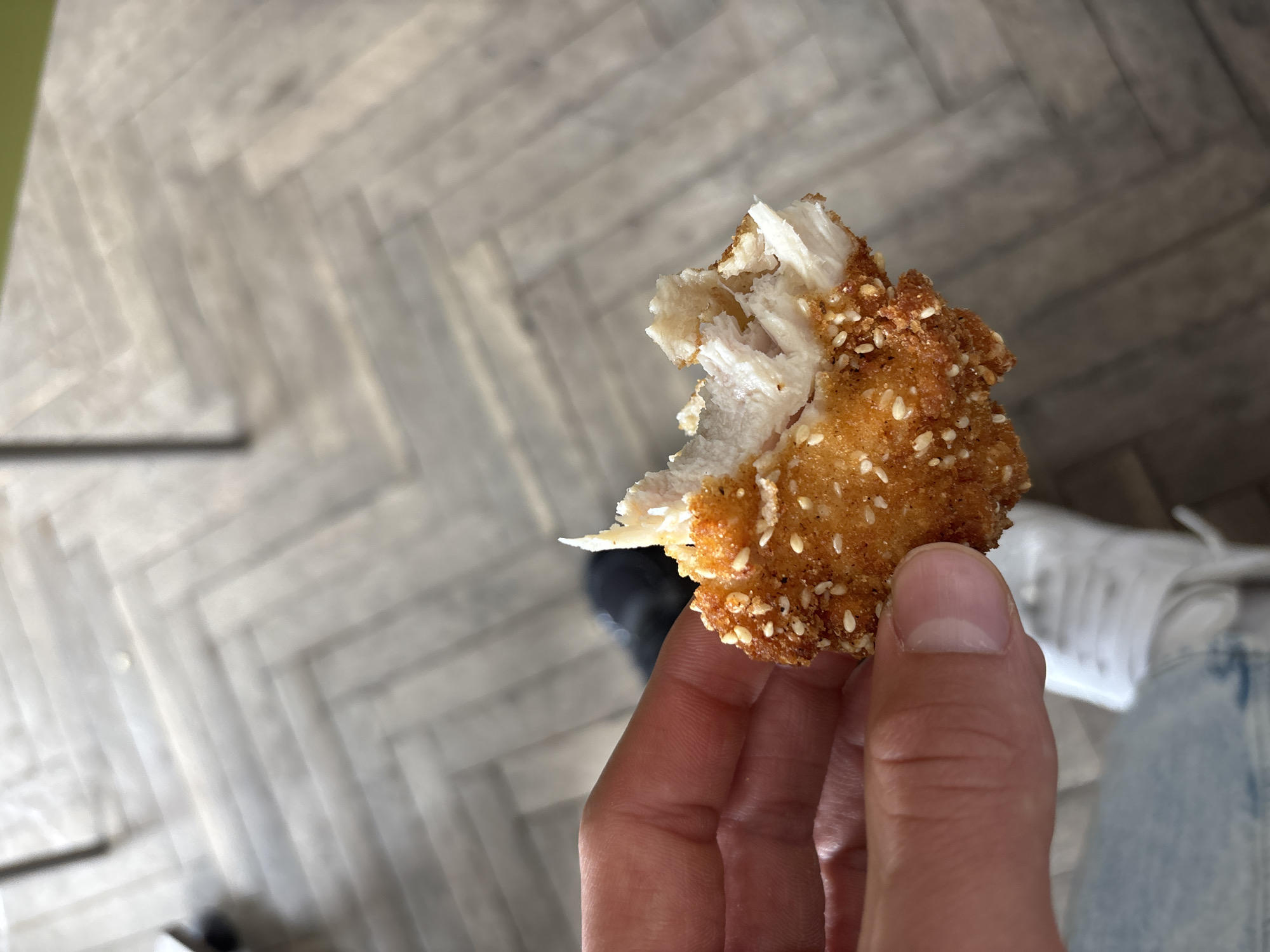A person holds a partially eaten, sesame seed-encrusted fried food item, likely chicken or a similar protein. The interior of the food item is white and fibrous, indicating cooked meat. The background is blurred, but shows a parquet-patterned floor and a glimpse of what appears to be a white shoe or sneaker, light blue jeans, and a dark-colored shoe. Based on the prompt's request, the location is Zlatograd, Bulgaria, though this is not visually evident. No specific interactions are apparent beyond the person holding the food.