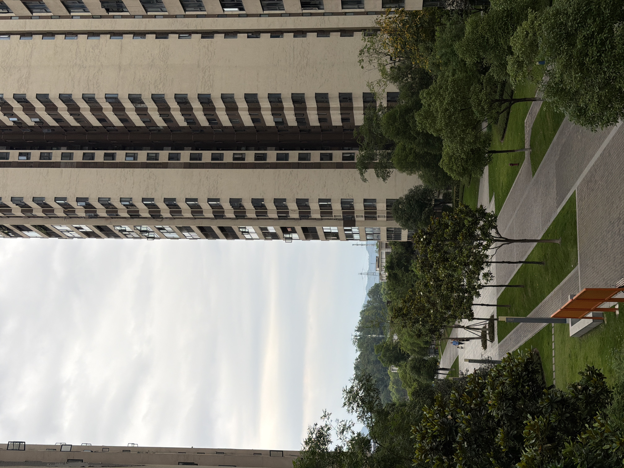 The image shows a view from an elevated position in the Chongqing High-tech Zone, China. Two tall buildings, each with numerous windows arranged in horizontal rows, dominate the top and bottom of the frame. Between them, a view opens to a cloudy sky and a green hillside with power lines. The foreground includes a park-like area with trees lining paved walkways, green lawns, and some park furniture. No people are clearly visible.
