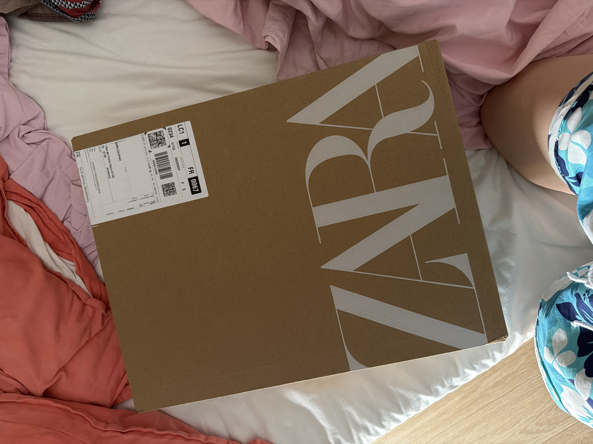 This media file shows a Zara package box resting on a white duvet, surrounded by a collection of pink and coral-colored fabrics, possibly bedding or clothing. The box is made of brown cardboard with the large, white "ZARA" logo prominently displayed across it. A shipping label is affixed to the upper left corner of the box, containing text in both French and English. The label includes the text "LC1 0234 FR 0067", "67100", "Z8Q30W2P", "ES4000 03 08.06.2025 21:57:200.30 kg 001/001 RTG 08062026 F2.000", "Izolda Gurchiani", "1 Rue des Frères Eberts 67100 Strasbourg", and "GLS service flexdelivery". The lower right portion of the image features a person's leg and hip, clad in blue and white floral patterned shorts. The overall scene suggests a casual, indoor setting, possibly a bedroom, with the focus on the arrival of a package. The lighting implies it is daytime.
