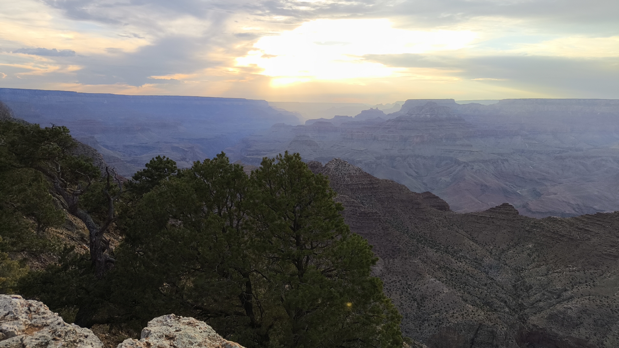 The image shows a panoramic view of the Grand Canyon at sunset. The sky is filled with dramatic clouds, with the sun breaking through in the distance, casting rays of light across the canyon. The layers of rock formations are visible, with shades of purple, brown, and red. In the foreground, there are pine trees and rocks, typical of the Grand Canyon landscape. The image appears to be taken from a viewpoint overlooking the vast expanse of the canyon, likely within Grand Canyon Village, Arizona, United States, known for its stunning viewpoints of the natural wonder. There are no people or animals visible in the frame.