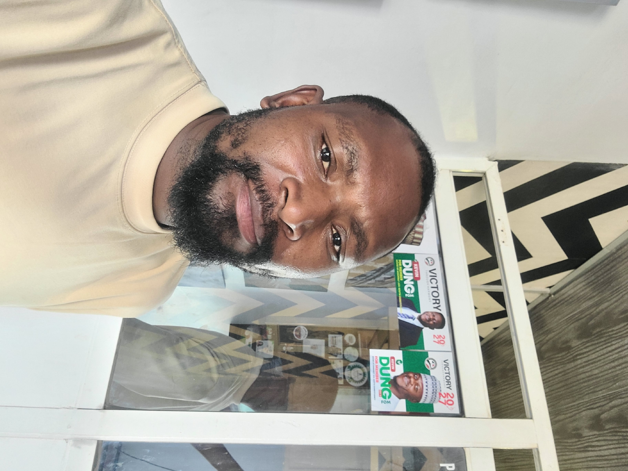 A mid-shot, selfie-style photograph captures a dark-skinned man with a beard, wearing a light tan, short-sleeved shirt. He is positioned in the foreground, looking directly at the camera. Behind him, to the right, is a white framed window with multiple posters visible. These posters feature pictures of men, text, and political campaign material. One poster prominently displays "VICTORY" and the name "KEVIN DUNG! FOR MEMBER." The background features a wall with a black and white geometric pattern and a wooden ceiling. The overall setting suggests an outdoor or semi-outdoor location, possibly a shopfront or a stall. The image is likely taken in Jos, Nigeria, given the context of political campaign posters for a local election. The lighting is natural and appears to be daylight.