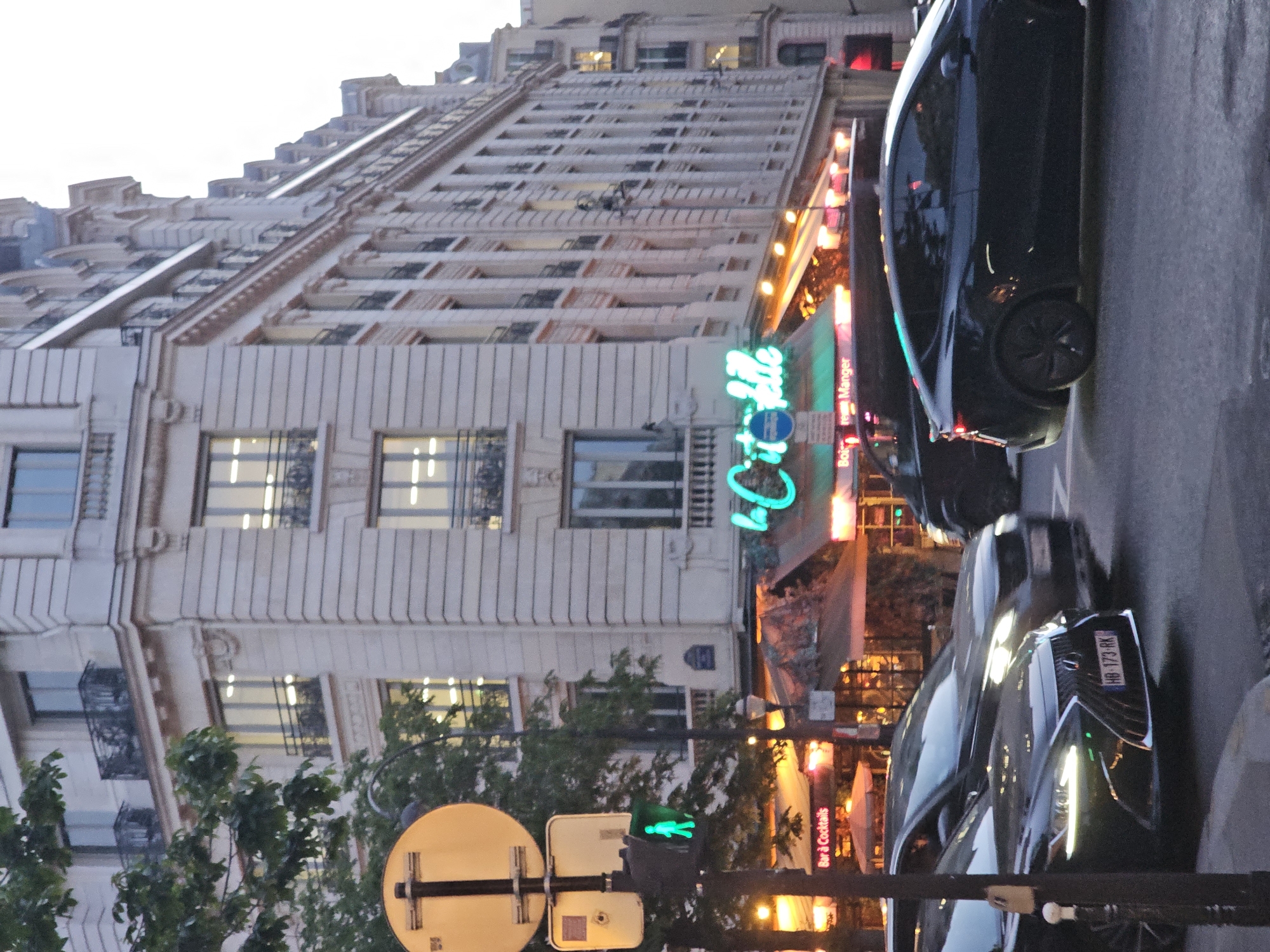 The image depicts a street scene in Paris, France, at dusk or twilight. The primary subject is a building of Parisian architecture, characterized by its light-colored facade, multiple stories, and rows of windows with ornate metalwork. A neon sign reading "Bobo Belle" indicates a restaurant or establishment at street level. Pedestrian traffic light signal and street signs are visible. Modern cars are parked along the curb.