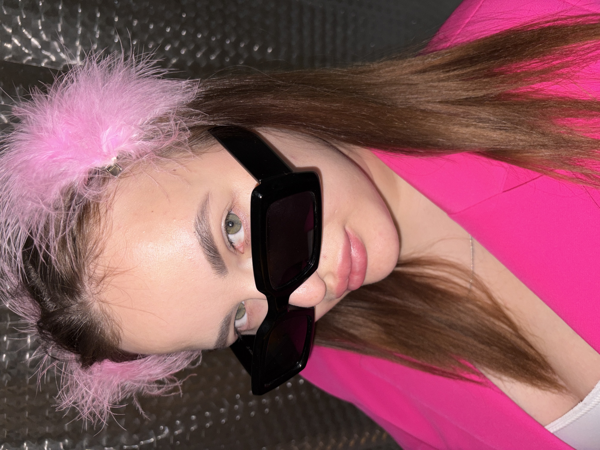 A young woman with brown hair and green eyes is posing for a close-up, slightly angled portrait. She is wearing black sunglasses and a pink feathered hair accessory. She's dressed in a bright pink blazer over a white top. The background appears to be a metallic or reflective surface. There is no evidence in the image to determine that the location is in the city of Krasnodar, Russia.