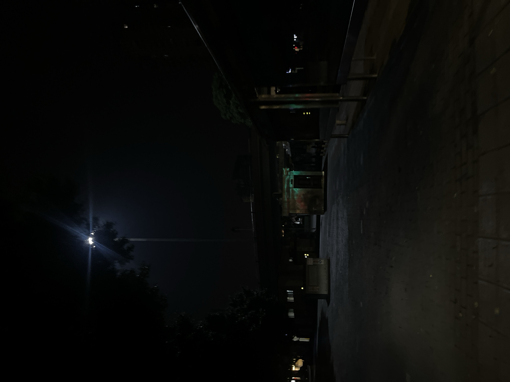 The image depicts a desolate nighttime urban environment in Jiulongpo District, China, illuminated primarily by a single intense overhead streetlight positioned centrally towards the upper frame, emitting significant lens flare. A large, dark tree silhouette occupies the left foreground, its foliage barely discernible against the dark sky. In the mid-ground, a concrete elevated structure, likely a bridge or light rail track, traverses horizontally across the scene, supported by prominent concrete pillars. Below this elevated structure, a building with several illuminated windows is visible on the left, while a small, shuttered kiosk-like structure stands prominently in the foreground. To the right of the kiosk, another structure emits a distinct green light. Along the right side of the frame, a covered walkway or platform with multiple vertical metallic supports extends into the distance. The ground consists of a paved surface, exhibiting variations in texture and shade, suggesting a mix of concrete and possibly tiled areas. Distant, faint artificial lights are visible further down the path on the right and across the background, indicating additional urban infrastructure. No individuals are present, and no discernable activity is occurring within the frame. The overall scene is characterized by deep shadows and high contrast due to the singular bright light source.