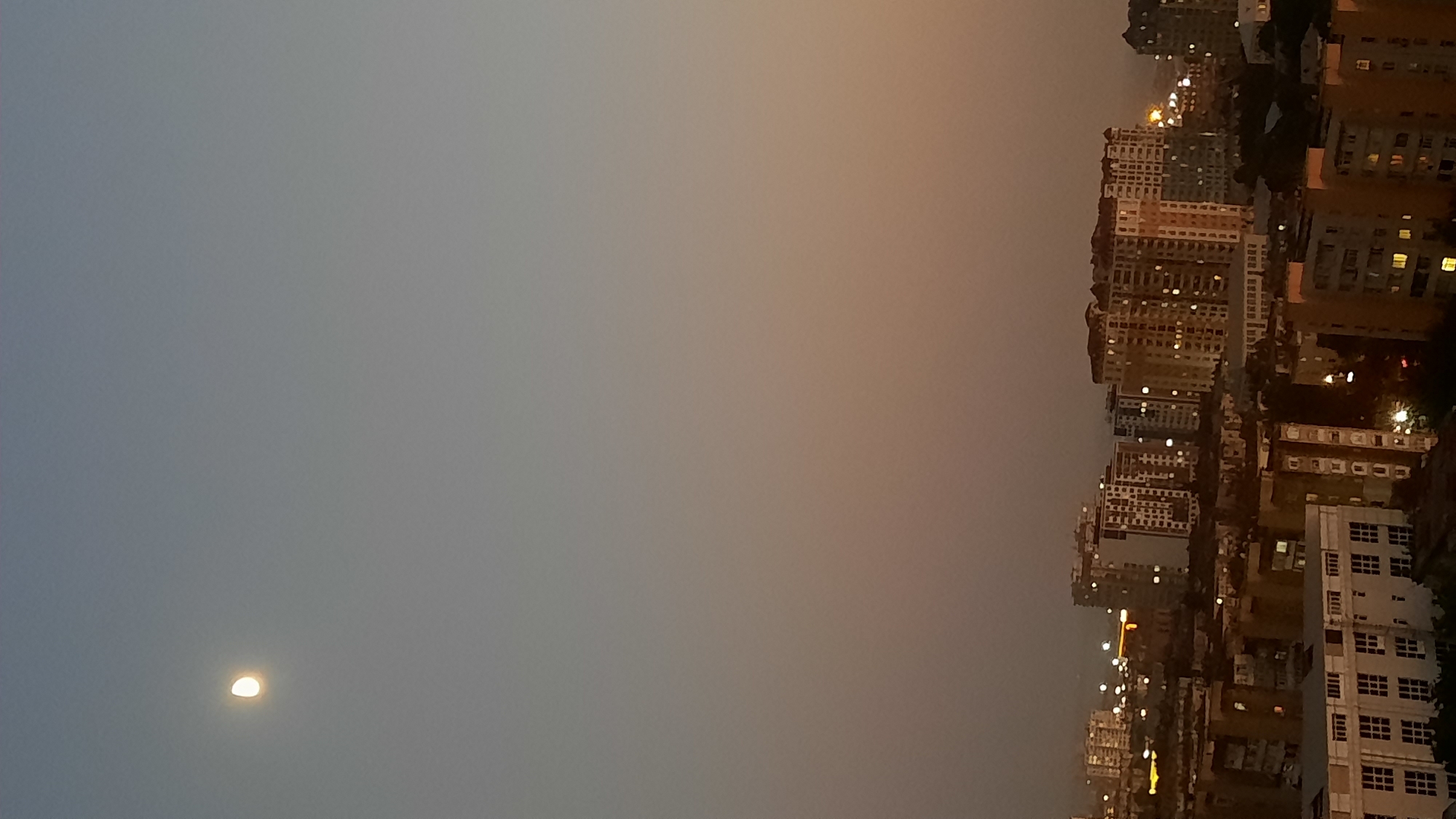 A vertical, slightly blurry, full shot captures a city skyline at dusk. The sky transitions from a light blue at the top to a warm orange-brown near the horizon, with the moon visible in the upper left quadrant. A dense collection of apartment buildings dominates the lower half of the frame, many with illuminated windows suggesting occupancy. The architecture varies, with some buildings appearing more modern with extensive glass, while others are older and more monolithic. The overall impression is of a residential area at the end of the day, with the moon overseeing the urban landscape.