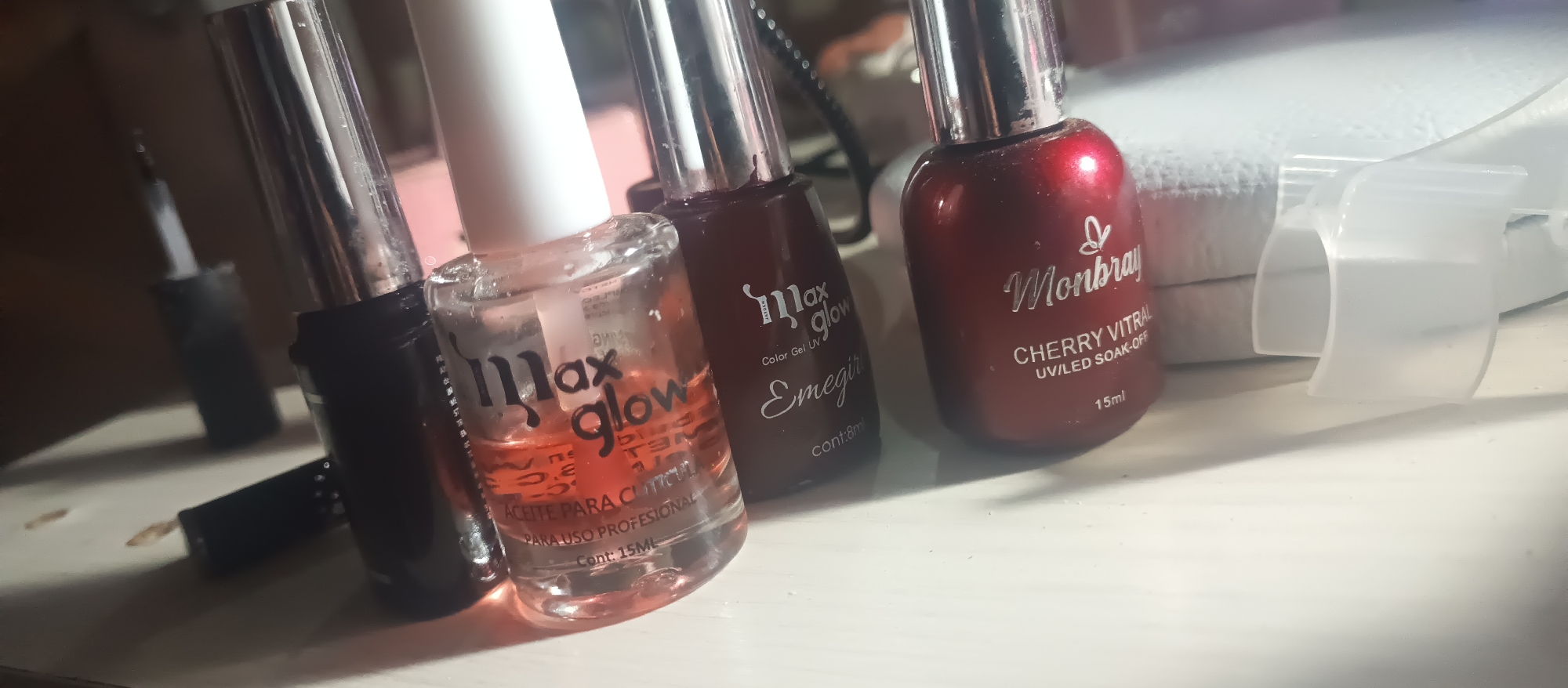In a close-up, dimly lit shot, several bottles of nail polish and related products are arranged on a white surface. From left to right, the visible items include a black nail polish bottle, a clear bottle labeled "max glow ACEITE PARA CUTICULA CONT: 15ML" which appears to contain an orangish liquid, a dark red nail polish bottle labeled "max glow Color Gel UV Emegirl cont: 8ml", and a bright red nail polish bottle labeled "Monbrary CHERRY VITRAL UV/LED SOAK-OFF 15ml". A white plastic object, possibly a nail drying accessory, is partially visible in the upper right corner of the frame. The background is blurred, suggesting an indoor setting, with indistinct shapes and colors. The lighting casts shadows and highlights on the bottles, creating a somewhat dramatic effect. There are no people or animals present in the image.