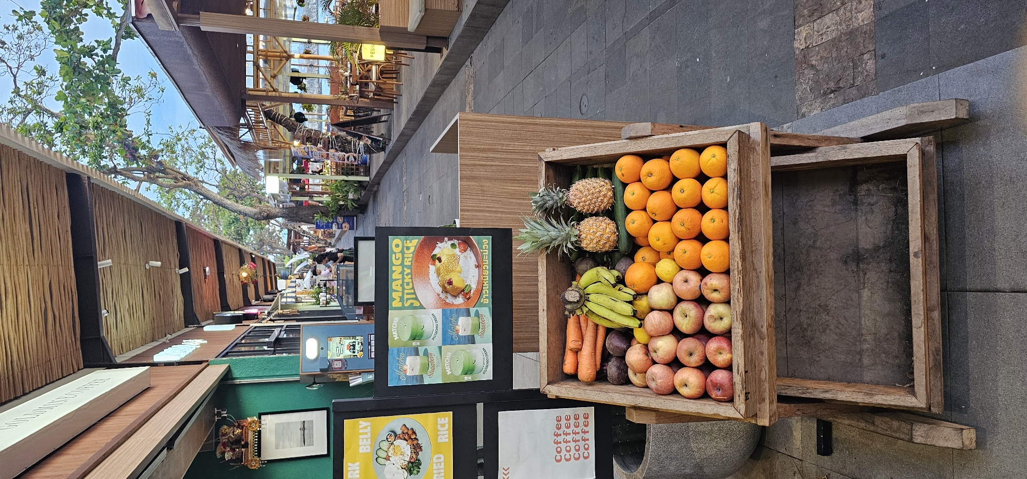 This vertically oriented image captures a lively outdoor market or food court scene in Denpasar, Indonesia, under bright daylight conditions.
In the foreground on the right, a multi-tiered wooden crate prominently displays an array of fresh fruits and vegetables. The top tier holds two pineapples, several green cucumbers, and numerous bright orange oranges. The middle tier features bunches of yellow bananas, carrots, and dark purple root vegetables, alongside some smaller yellow fruits. The bottom tier is filled with red and green apples. Adjacent to this full crate, an empty wooden frame stands on a paved surface, with a grey planter visible beneath the fruit display.
To the left of the fruit display, several dark signs advertise various food and drink offerings. A large sign prominently features "MANGO STICKY RICE" with an enticing image of the dessert and its Thai translation, "ข้าวเหนียวมะม่วง." Below this, "Matcha" beverages are advertised, including "Pistachio Cream" and "Strawberry" flavors. Another partially visible sign shows "PORK BELLY FRIED RICE" with the text "RK BELLY," and a sign repeating "Coffee" is also visible.
The background reveals an expansive outdoor setting with a tiled walkway. On the left, a row of stalls with textured wooden facades, possibly made of bamboo or similar material, extends into the distance. Green trees with sprawling branches overhang these structures, providing natural shade. On the right side of the walkway, open-air dining areas with wooden tables and chairs can be seen under a wooden-beamed roof structure, with a sign showing "PESS." Several people are present, either walking along the path or seated at the establishments, indicating a bustling atmosphere. The scene suggests a vibrant culinary destination offering both fresh produce and prepared dishes.