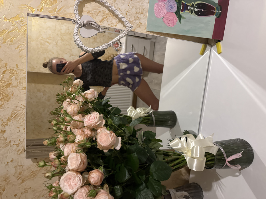 A mirror selfie captures a young woman in a cropped top and patterned shorts. She holds a phone, her face obscured. A large bouquet of pale pink roses dominates the foreground, obscuring the view. Behind her is a white heart-shaped decoration on the mirror and a painting of flowers in a vase mounted on a white cabinet. The background suggests a neutral-toned wall. The bouquet is tied with white ribbons.