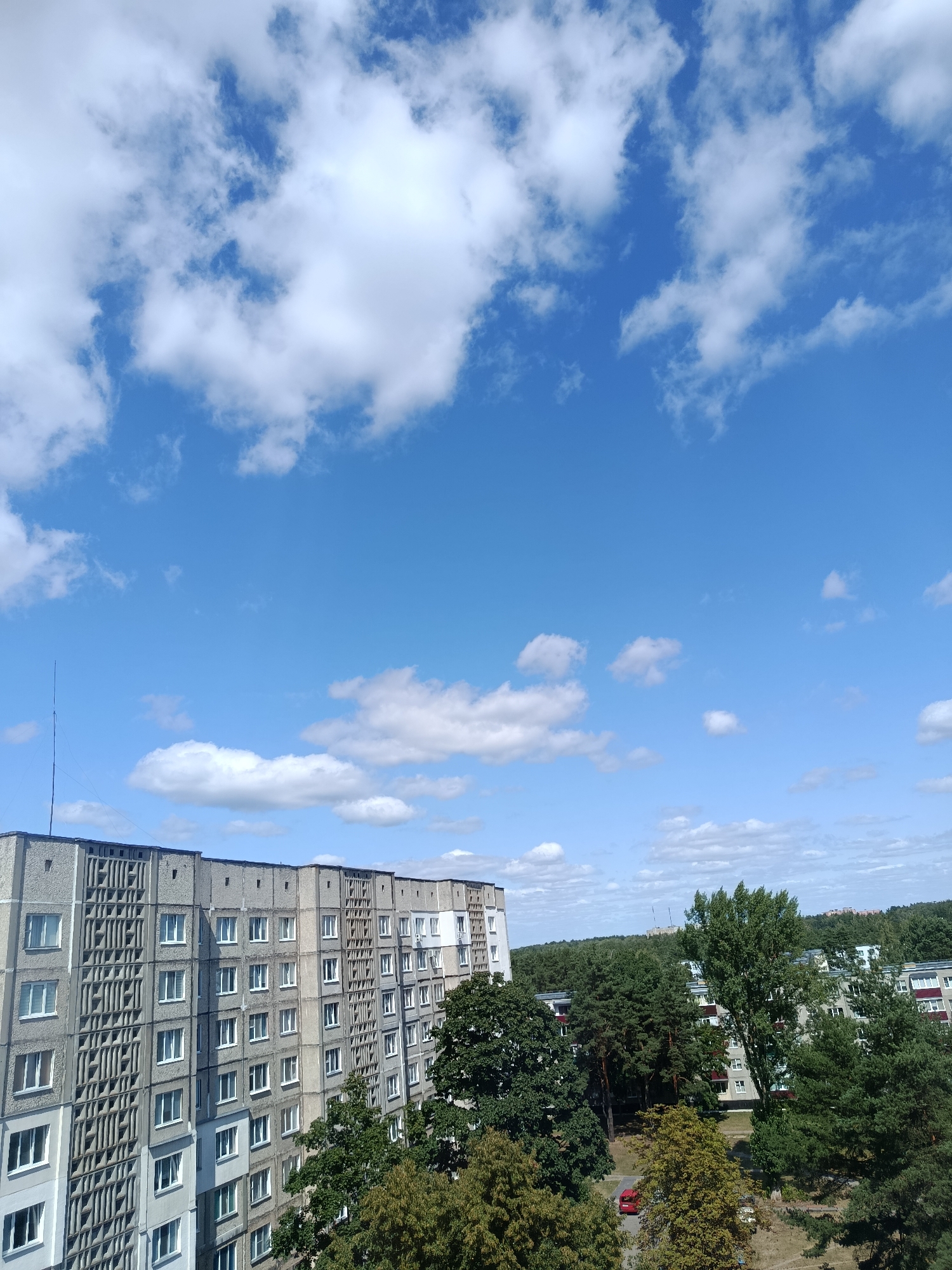 The image shows a view of a Soviet-era apartment building in Svietlahorsk, Belarus, against a bright blue sky with scattered white clouds. The building is tall and multi-story, with a repetitive facade of windows and concrete paneling. Dense green trees surround the building, some of which are tall evergreens, while others are deciduous. The trees partially obscure other residential buildings in the background. A red car is visible in the foreground, parked on a patch of ground between the trees and the apartment building. The scene appears to be an outdoor urban environment on a clear day.