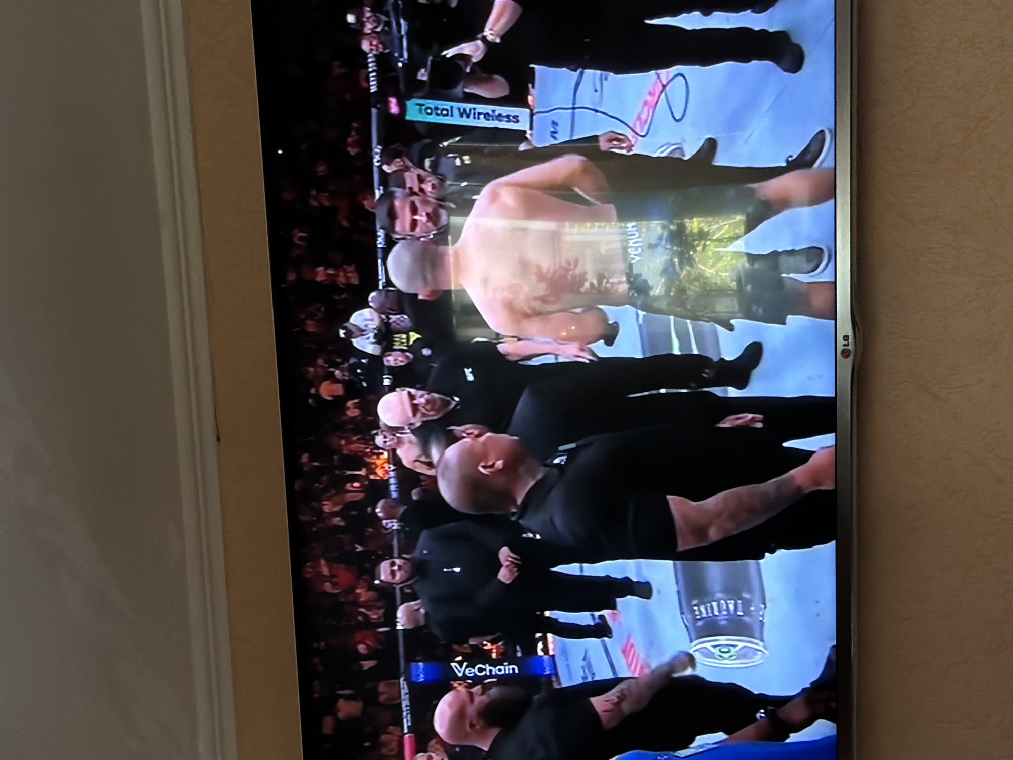 The image shows a scene from a televised UFC event. A shirtless male fighter with visible tattoos on his back is surrounded by security personnel in black suits. The fighter is wearing shorts with "VENUM" branding. The event is taking place in a ring with advertisements for "Total Wireless" and "VeChain" on the mat. The scene includes a crowd of spectators in the background. There is no information about location in the city of Beryozovka, Russia.