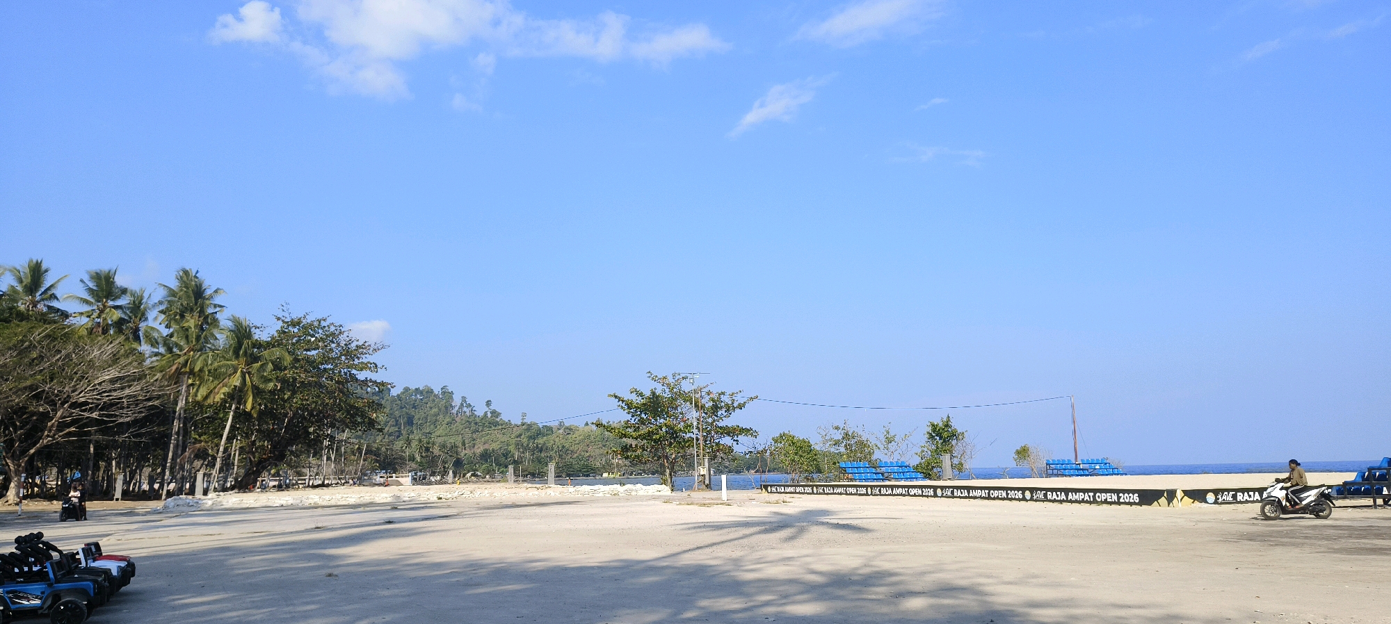 The image depicts a wide, sandy area in front of a beach in Waisai, Indonesia. The sky is a clear, bright blue with a few wispy clouds. Palm trees and other lush vegetation line the background, with a rolling, tree-covered hill visible further in the distance. In the middle ground, there is a section of beach with blue stadium seating and a fence with text that reads "RAJA AMPAT OPEN 2026". The sandy foreground is mostly empty, with a few small toy vehicles parked on the left and a person riding a scooter on the right. The overall impression is a tropical coastal location under a sunny sky.