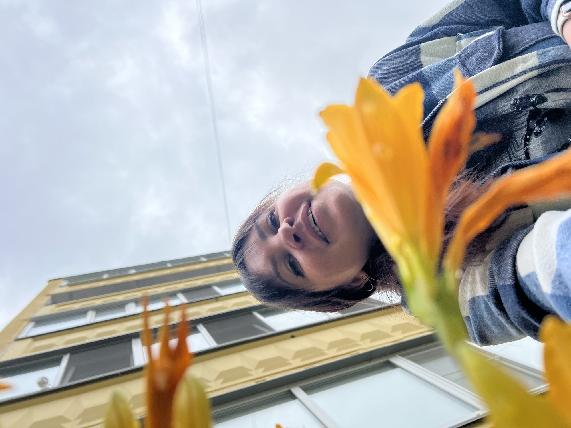 From a low-angle perspective, a woman's face is framed by orange-yellow flowers. She has brown hair and is wearing a blue and white plaid shirt. Behind her, a building with multiple floors and white-framed windows rises into the sky. The sky is overcast. The scene is in Noviy, Russia.