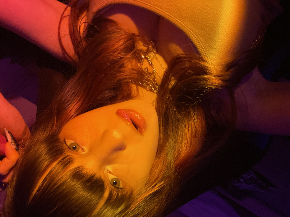 A close-up, angled portrait depicts an individual in an indoor setting in Raanana, Israel. The subject, appearing female, is positioned with the head tilted and eyes, appearing light green or hazel, gazing directly forward. Long, reddish-brown hair with bangs covers the forehead and frames the face; fine strands of glitter or tinsel are visible within the hair. The subject's lips are glossy, colored light red, with the tongue slightly extended to touch the upper lip. A light-colored, sleeveless garment is worn, and multiple layered necklaces, including one with a Star of David charm, adorn the neck. A hand, with dark-painted fingernails and visible white patterns, is partially visible at the top of the frame, holding a small, red, spherical object near the subject's head. The scene is illuminated by dominant artificial orange and red light, with areas of purple light present in the dark background, suggesting a low-light environment.