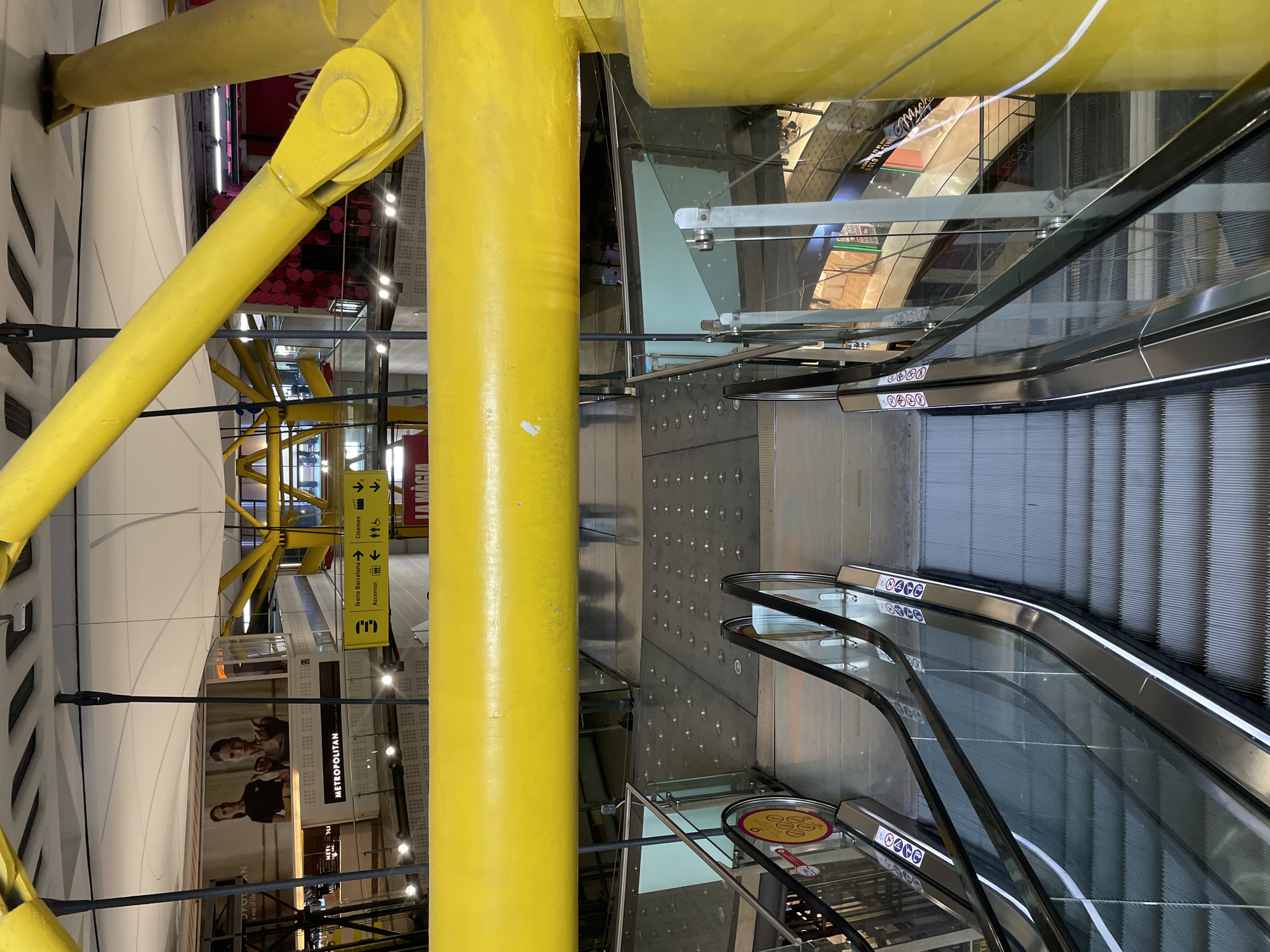 This media file depicts the interior of a brightly lit, modern multi-story building, likely a shopping mall or large commercial complex, given the visible signage and escalators. The dominant features are several massive, bright yellow structural beams and columns that crisscross the frame, adding a bold architectural element.
On the right side of the image, two escalators descend to lower levels. They feature transparent glass balustrades and sleek black handrails. Numerous white circular safety signs with red borders and black pictograms are visible on the escalator railings, indicating rules such as holding the handrail, no running, and restrictions on certain items or bare feet.
The building's interior includes multiple levels connected by these escalators and possibly elevators, as indicated by signage. Glass barriers and railings are used throughout, offering views of different sections of the complex. A wall to the right of the central yellow column features a textured surface with many small, circular, illuminated elements.
Visible text provides context for the location and establishments within. A sign in the mid-ground points to "Ikono Barcelona" with a left arrow, and "Cinemes" (cinemas) and "Ascensor" (elevator) with arrows pointing up and right, confirming the location as Barcelona, Spain. Another prominent sign reads "METROPOLITAN" with "a way of life" below it, likely for a gym or lifestyle brand. Other partial texts include "LA MÀCIA," "Mich," and "POP."
No individuals are distinctly visible, presenting an uncrowded atmosphere. The consistent artificial lighting suggests an indoor setting, and no specific time of day or weather conditions can be inferred. The overall impression is one of a clean, well-maintained, and architecturally striking commercial environment.