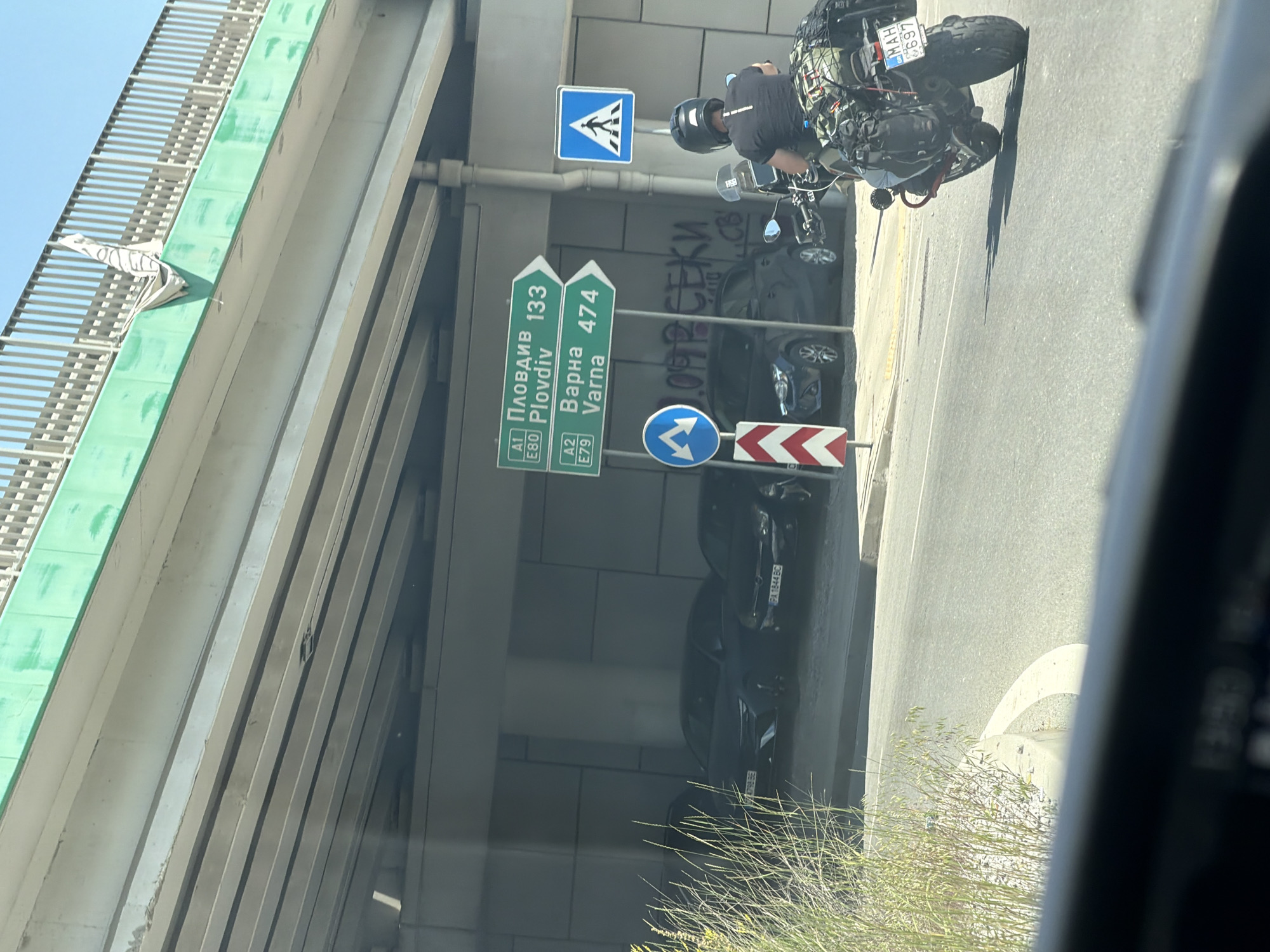 The image shows a view from a vehicle on a sunny day, looking towards a multi-level road infrastructure. In the foreground, the top of a car's dashboard is visible, with a blurry reflection. A motorcycle with a rider wearing a helmet and carrying luggage is in the middle ground, moving away from the camera. The rider is wearing a dark shirt and jeans, and the motorcycle is equipped with multiple bags.
Behind the motorcycle, several cars are parked under an elevated roadway. The cars are dark in color, and some license plates are partially visible. One license plate reads "MAH 697".
Road signs are prominent in the scene. Two green directional signs are mounted on poles. The top sign indicates "A1 Plovdiv 133" and "E80 Plovdiv 133". The bottom sign shows "A2 Varna 474" and "E79 Varna 474". A blue square sign with a white pedestrian symbol is also visible, indicating a pedestrian crossing. A circular blue sign with two white arrows pointing left and right, and a red and white striped marker are also present.
Graffiti in Cyrillic letters is visible on a concrete wall in the background. The overall environment appears to be an urban setting with significant road construction or infrastructure. The location is identified as Sofia, Bulgaria, based on the Cyrillic text and the road signs which include Bulgarian destinations.