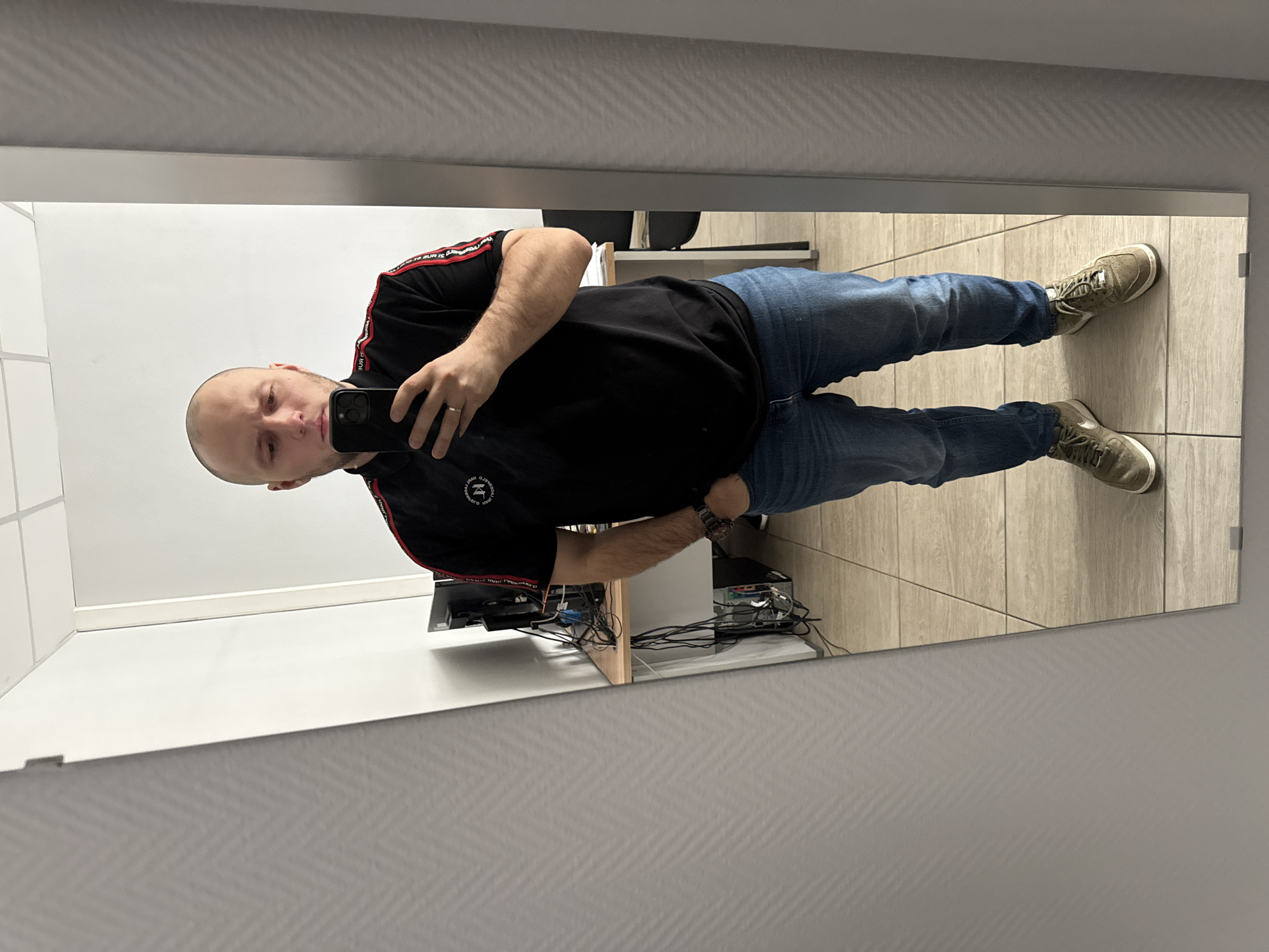 A bald man wearing a black shirt with red and white stripes on the sleeves, blue jeans, and olive green sneakers is taking a selfie in a mirror. He is holding a black smartphone in his left hand. A silver ring is visible on his left ring finger. Behind him, a desk or shelf is visible with several electronic devices and wires, and a black office chair. The floor is tiled with light brown rectangular tiles. The walls appear to be white and grey with a subtle texture. It is not possible to determine the location specifically in Krasnoyarsk, Russia, based on the image.