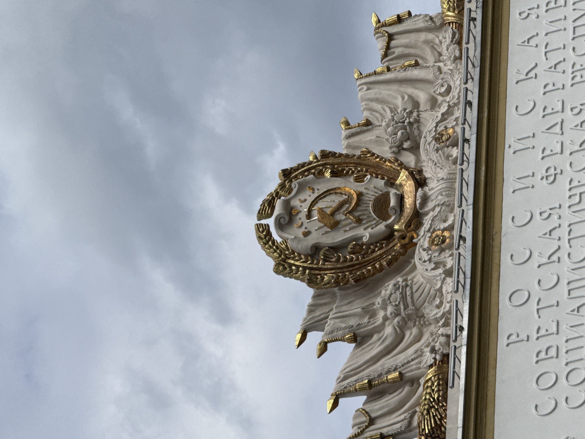The image shows a close-up view of a building facade in Moscow, Russia, under a cloudy sky. The facade features ornate architectural elements, including a large golden emblem with the hammer and sickle symbol, representative of the Soviet Union. The emblem is surrounded by draped fabric-like sculptures and golden tassels. Below this, carved into the white stone of the building, are Cyrillic letters. The visible text reads "РОССИЙСКАЯ СОВЕТСКАЯ ФЕДЕРАТИВНАЯ СОЦИАЛИСТИЧЕСКАЯ РЕСПУБЛИКА," which translates to "Russian Soviet Federative Socialist Republic." This inscription, along with the emblem, strongly suggests the building is a monument or structure associated with the Soviet era, possibly located at the VDNKh (Exhibition of Achievements of National Economy) park, which is known for its Soviet-era pavilions and monuments. There are no people visible in the image.