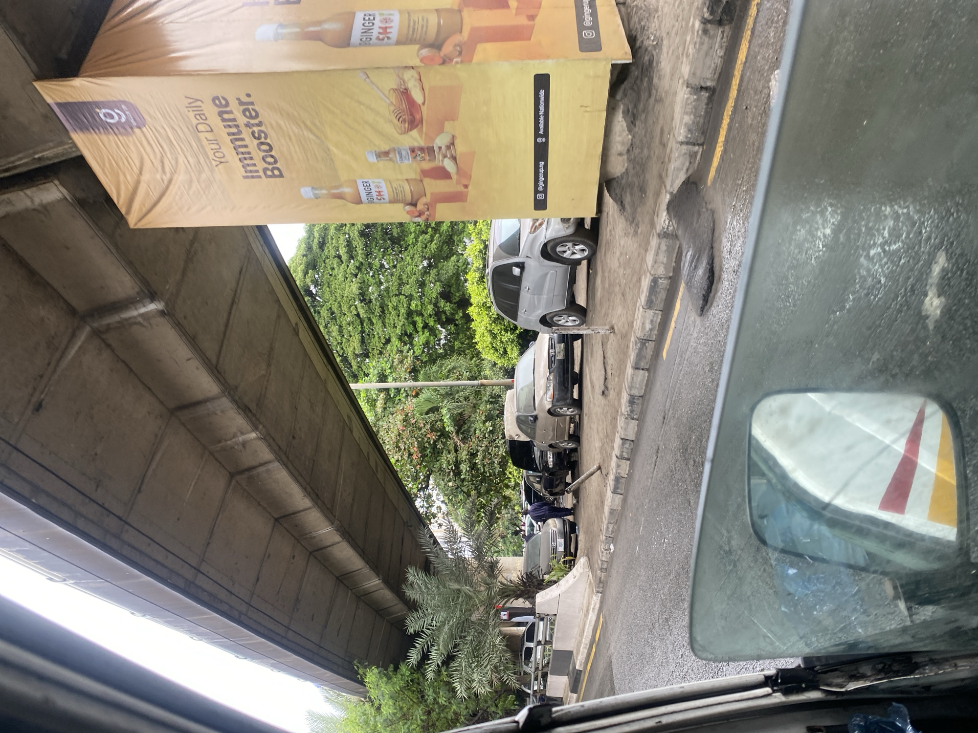The image depicts a street scene in Victoria Island, Nigeria, beneath an elevated concrete structure, likely a highway overpass. A large, yellow billboard advertising "Your Daily Immune Booster" is prominently displayed on the right side of the frame. Several cars are parked along the side of the road, including a silver SUV and a beige sedan. A person in a blue uniform is standing near a black car. Trees and vegetation are visible behind the parked cars and under the overpass. The road surface appears wet and there is a pile of dark debris on the side of the road. The photo is taken from inside a vehicle, as evidenced by the reflection in the side mirror.