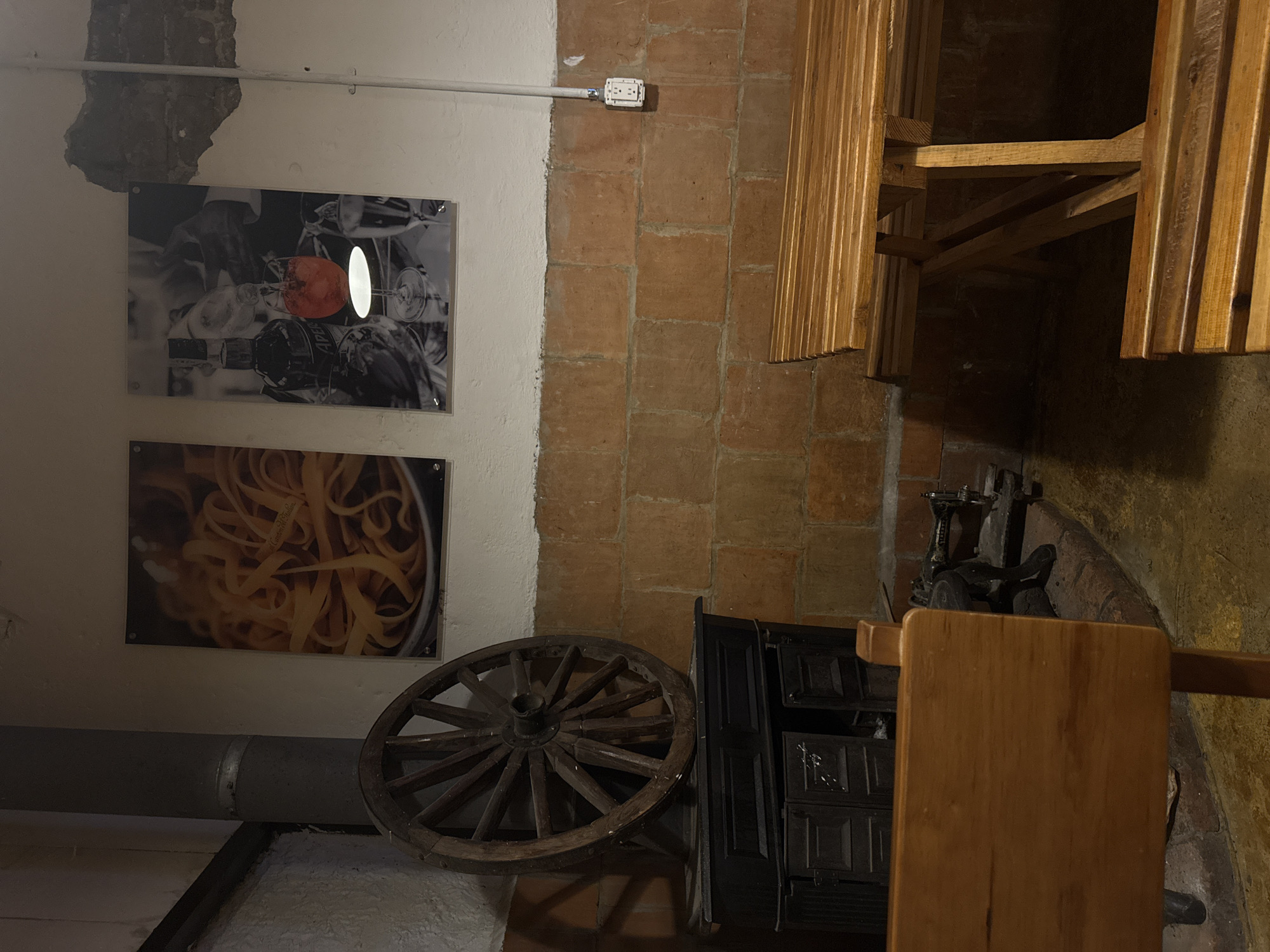 The image depicts an interior scene, possibly a restaurant or a decorative space in Chipilo de Francisco Javier Mina (Chipilo), Mexico. On the left wall are two framed pictures: one featuring a still life with what appears to be wine glasses and a red object, and another showcasing pasta. Below the pasta picture, a gray pipe is visible against the wall.
The right side of the image shows a brown brick wall. Adjacent to the brick wall is a wooden slat structure, potentially part of a seating arrangement. A dark-colored cabinet stands next to a wooden wheel. On top of the cabinet, there is an old-fashioned appliance, possibly a grinder, and a portion of a wooden table. The floor appears to be uneven and made of a textured material.