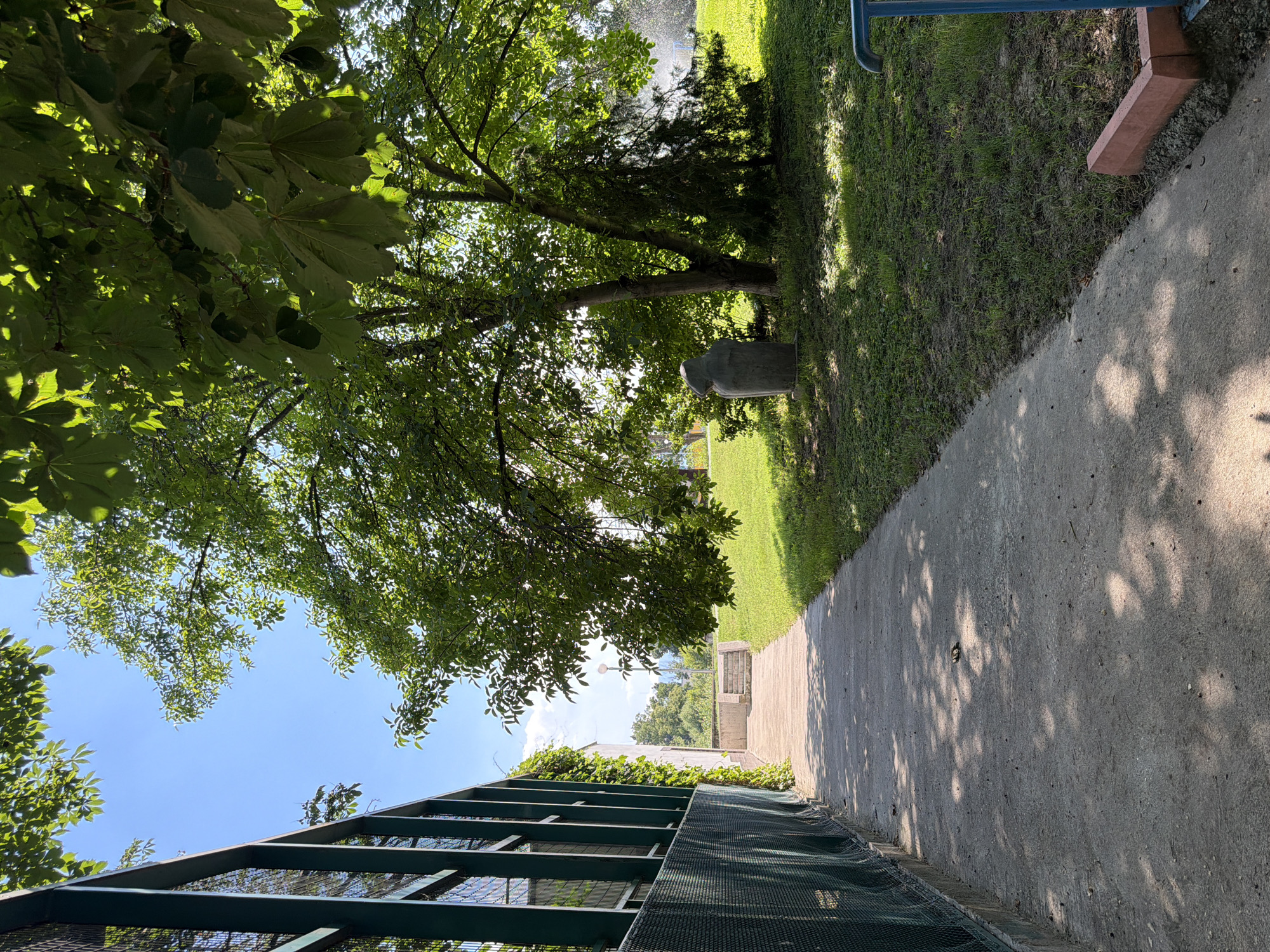 The image shows a pathway in an outdoor setting, likely a park or a recreational area, in Pazardzhik, Bulgaria. A green metal fence runs along the left side of the pathway, casting shadows from the overhead foliage. The pathway itself is made of concrete and is partially shaded by the leaves of a large tree on the right. To the right of the pathway, a grassy area is visible, with a small stone sculpture positioned near the base of a tree. In the background, a set of concrete stairs leads up to a higher level, and beyond that, trees and buildings are visible under a clear blue sky. The overall scene depicts a sunny day in a tranquil outdoor space.