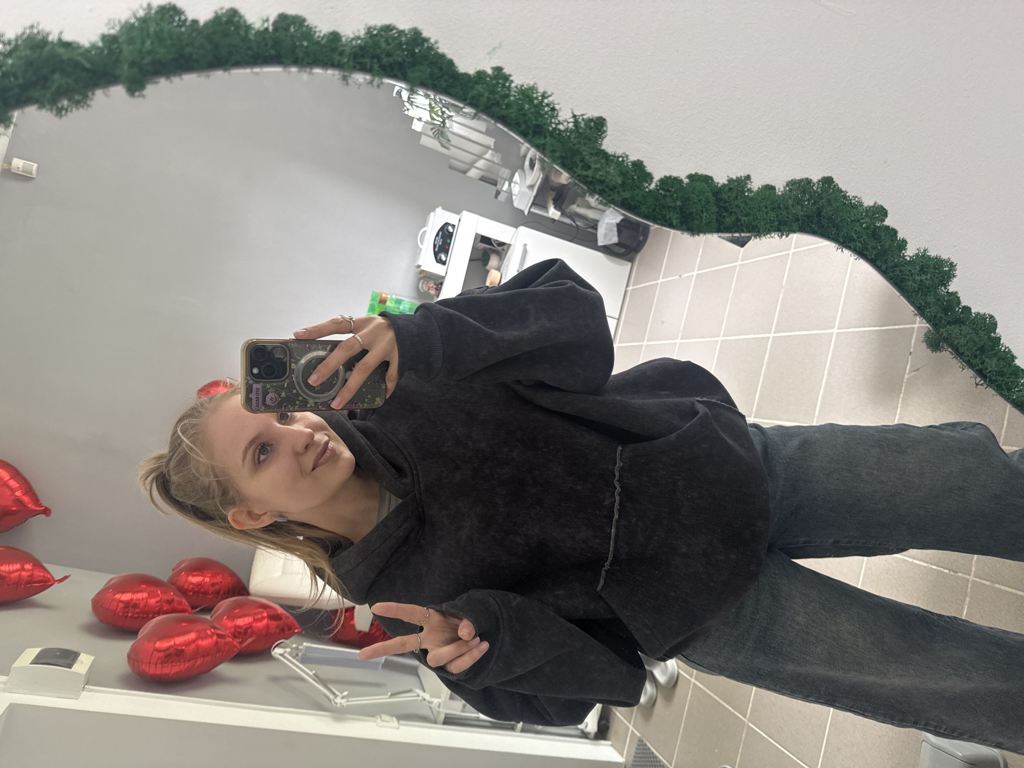 A young woman is taking a selfie in a mirror. The mirror is decorated with greenery around the edge. She is wearing a dark grey hoodie and dark grey jeans. She is making a peace sign with her left hand. The room appears to be a bathroom or small room with tiled floors. The reflection in the mirror shows a white cabinet with a microwave and other items on it. There are also several red heart-shaped balloons in the reflection. The location is Izhevsk, Russia.