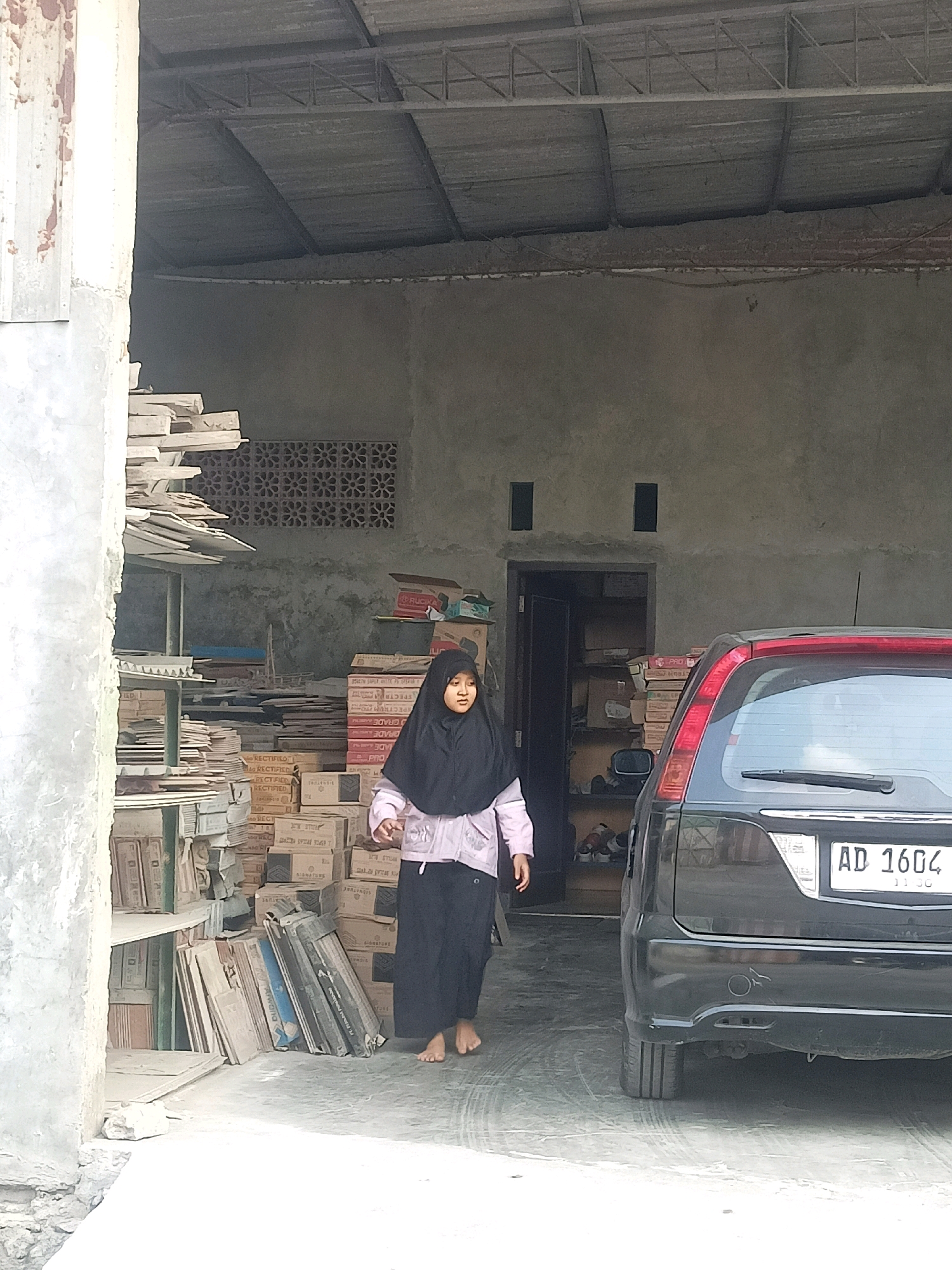 A young female wearing a black hijab and a light pink jacket over a black garment walks towards the viewer's left in a dimly lit, open-air storage area in Karangtalun, Indonesia. She is barefoot. To her right is the rear of a black SUV, bearing the license plate "AD 1604". The background consists of a concrete wall with two small, dark, rectangular openings. Stacks of boxes and what appear to be tiles are visible on shelves to the left and in piles behind the girl. The floor is concrete with visible markings. The scene is outdoors with a corrugated metal roof structure visible overhead.