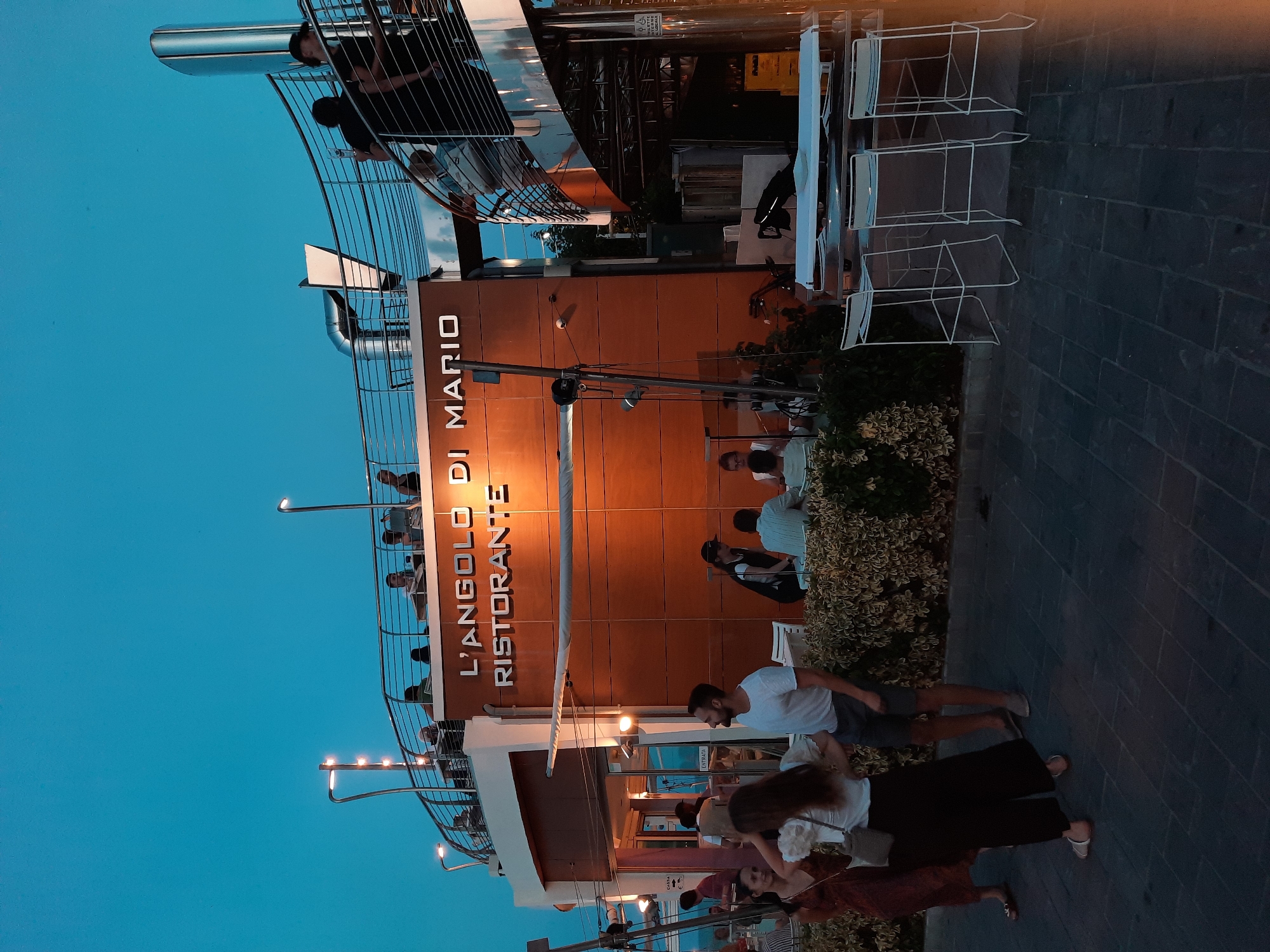The image depicts an exterior view of "L'ANGOLO DI MARIO RISTORANTE" in Pesaro, Italy, at dusk. A multi-level structure with an orange facade is visible, bearing the restaurant's name in white lettering. The upper level features a railing and several patrons seated at tables. A spiral staircase is visible on the right side of the building. In the foreground, several people are standing and walking on a paved area. White bar stools and a table are set up in front of the restaurant. The sky is a deep blue, indicating it is twilight.