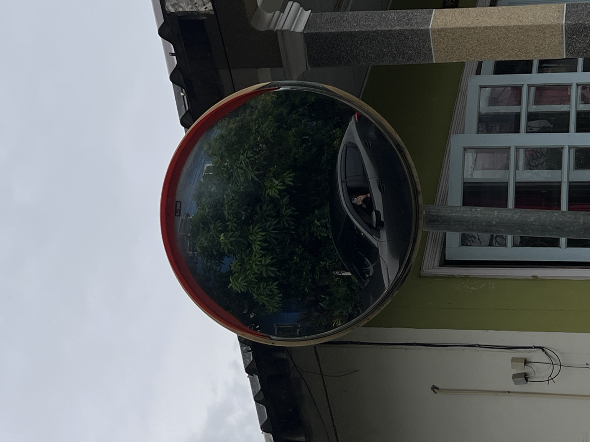 A large, round, convex traffic mirror with a reddish-orange rim is positioned prominently in the center of the image. The entire image is oriented vertically, with the sky on the left and a building structure on the right.
Inside the mirror's reflection, dense green foliage, likely trees or bushes, occupies the left and central parts. On the right side of the reflection, a dark-colored car, possibly an SUV or hatchback, is partially visible. A person's head and arm can be seen inside the car's window, appearing to hold a device, possibly a phone, indicating they are likely the photographer capturing their reflection.
Outside the mirror, the upper left area shows a grey, overcast sky. To the right of the mirror, a building wall painted a light green or lime green color is visible. A multi-paned window, framed in white or light blue, is set into this green wall, with glimpses of red fabric or objects inside. Above and to the left of the mirror, dark architectural elements, including a column or beam with a patterned texture and white trim, are present. Below the mirror, the dark, ridged eaves of a roof can be seen. Further down the building wall, a lighter-colored section is visible, with black electrical wires and two small white electrical boxes attached.
A small, blurry label on the reddish rim of the mirror appears to show "R 200" or similar text. The overcast sky suggests it is daytime. The presence of the mirror, car, and building indicates an urban or suburban setting, possibly near a driveway or street corner.
