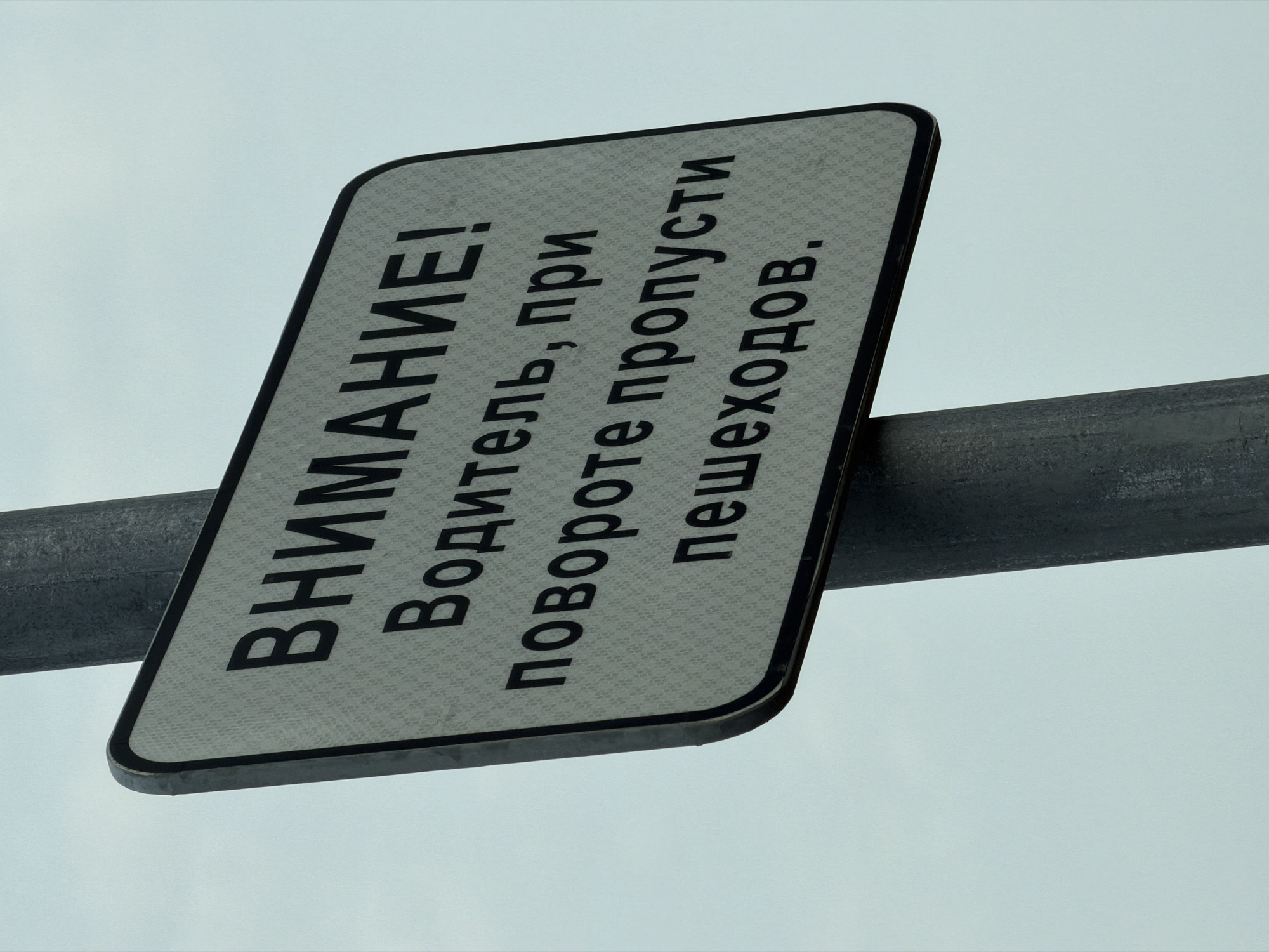 A road sign in the town of Поварёнки, Russia, displays a message in Russian: "ВНИМАНИЕ! Водитель, при повороте пропусти пешеходов." (Attention! Driver, yield to pedestrians when turning). The sign is rectangular with rounded corners, white with black lettering and a black border. It's mounted on a gray metal pole against a light gray sky.