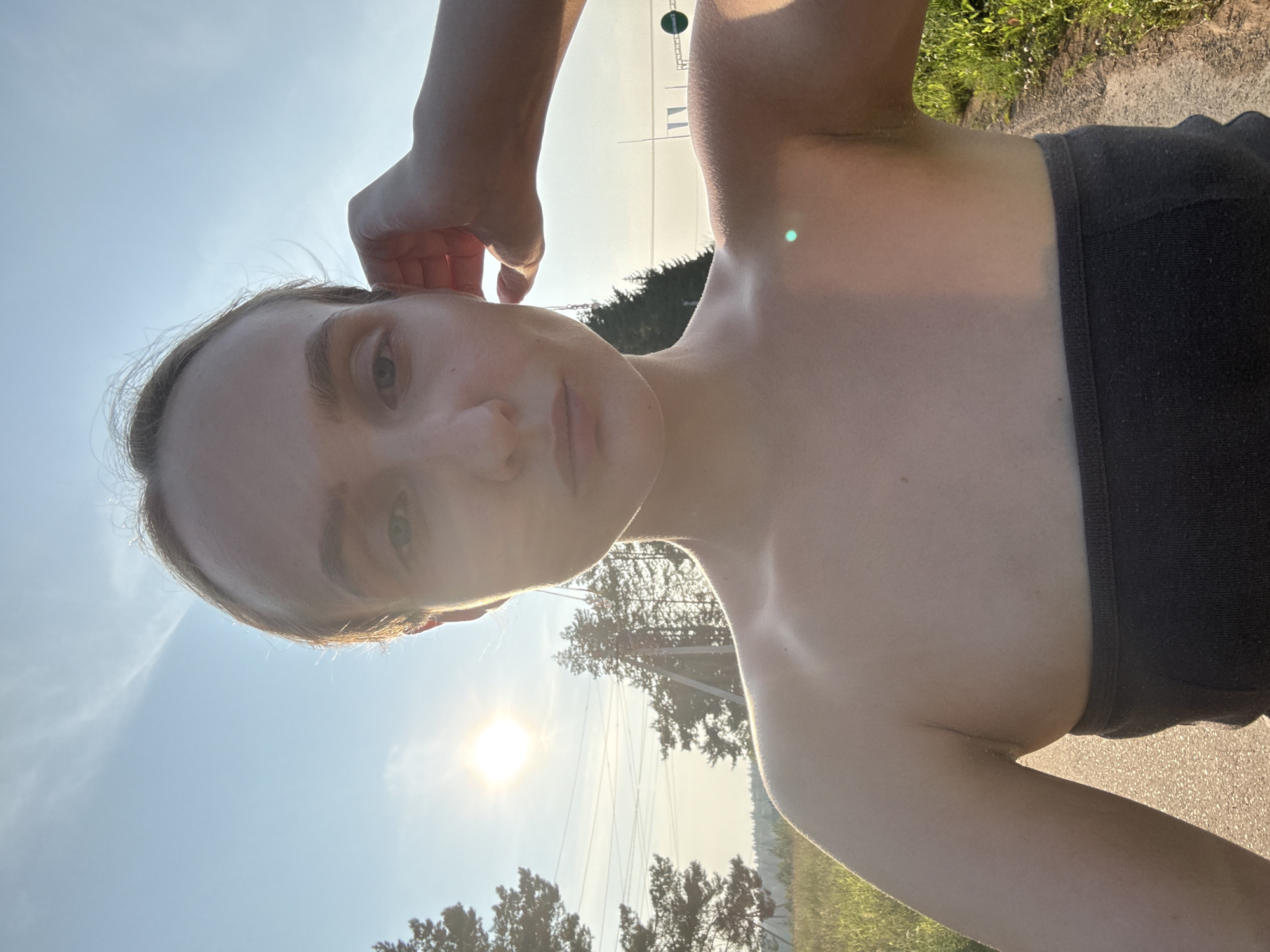 A young woman is captured in a selfie taken outdoors in the village of Podshivalovo, Russia. The sun is setting or rising, casting a warm glow across the scene. The woman is wearing a dark strapless top and has her right hand resting on her head. She is positioned on a dirt path, with tall grass and trees visible on either side. In the background, power lines and utility poles suggest the presence of infrastructure. The sky is bright blue with some wispy clouds.