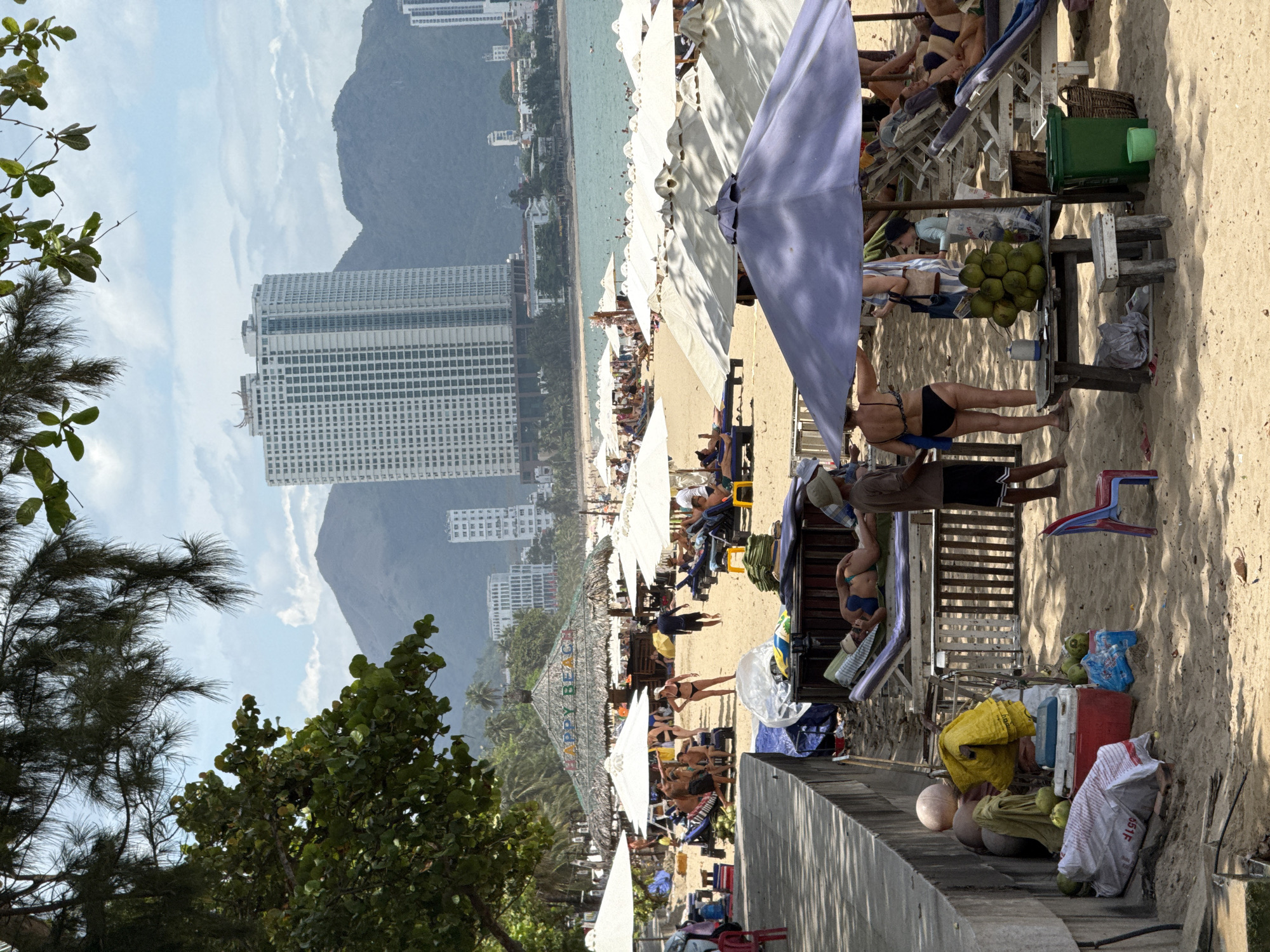 This image depicts a bustling beach scene on a sunny day in Nha Trang, Vietnam, likely near the main city beachfront. In the foreground, a concrete wall borders a sandy area filled with beach chairs, umbrellas, and various objects. Several people are present, some lounging on chairs, others walking or standing. A thatched-roof structure with the words "HAPPY BEACH" visible is a central point of interest, with umbrellas set up around it. In the background, the turquoise ocean stretches towards a hazy horizon, dotted with small boats and more beachgoers. Towering over the scene are modern, high-rise buildings, characteristic of Nha Trang's urban coastline, and lush green mountains rise behind them, creating a dramatic backdrop. The overall atmosphere is one of a popular tourist destination enjoying favorable weather.