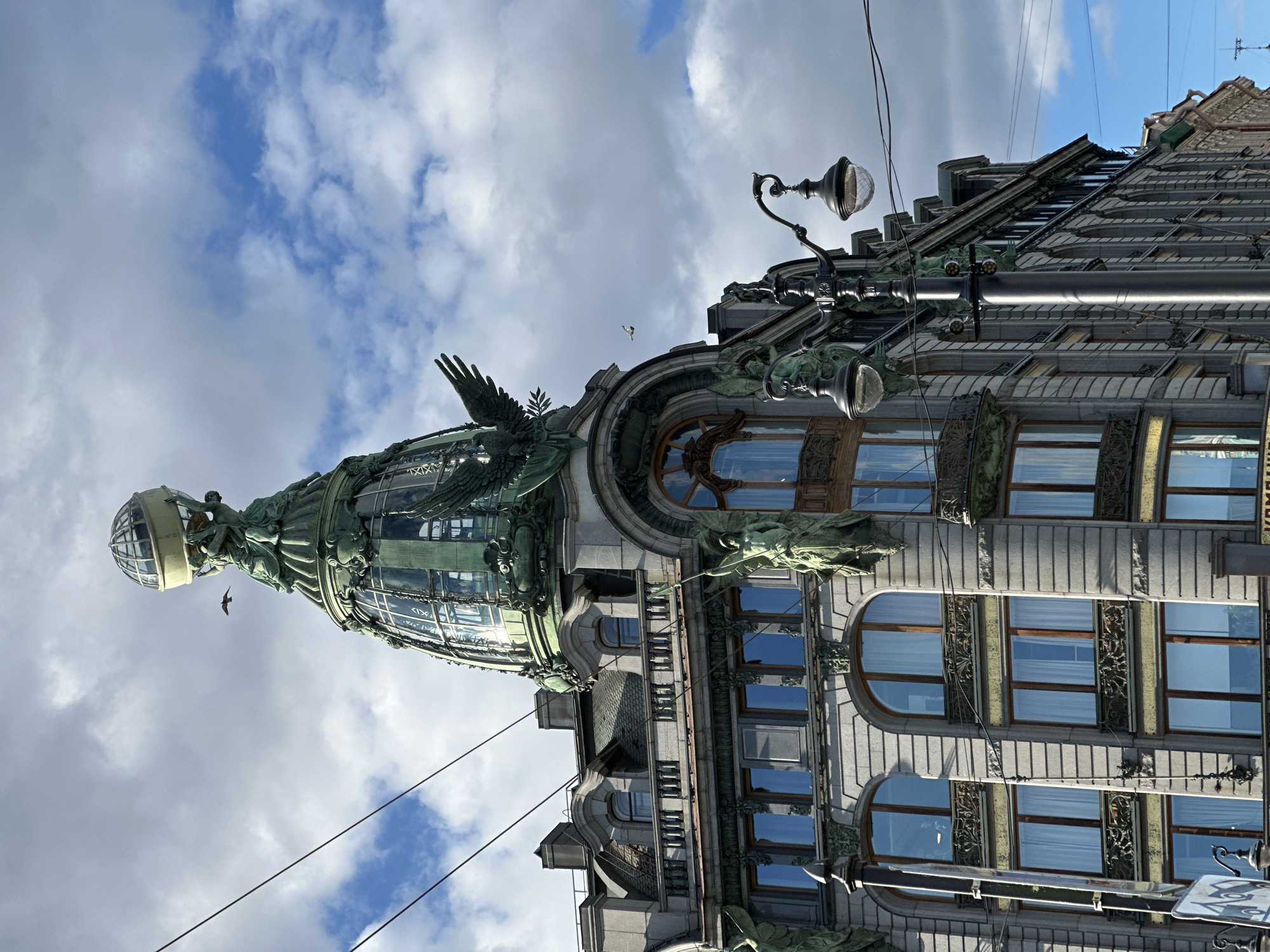 The image shows the Singer House, also known as the Dom Knigi (House of Books), in Saint Petersburg, Russia. It is an Art Nouveau building located on Nevsky Prospekt, a prominent street in the city. The photograph is taken from a low angle, looking up at the building's ornate facade and distinctive dome.
The dome, made of glass and metal, is topped with a large globe supported by sculptures of figures and eagles. The building itself is constructed of light-colored stone and features numerous windows, many of which are arched. Decorative elements, including sculptures of winged figures and relief carvings, adorn the exterior. A streetlamp with two globes stands prominently in the foreground on the right side of the image, casting shadows on the building.
The sky is partly cloudy, with patches of blue visible between the white clouds. Two birds are in flight, one near the top of the dome and another further away in the sky. Power lines are visible crisscrossing the frame. The overall impression is one of architectural grandeur and historical significance.